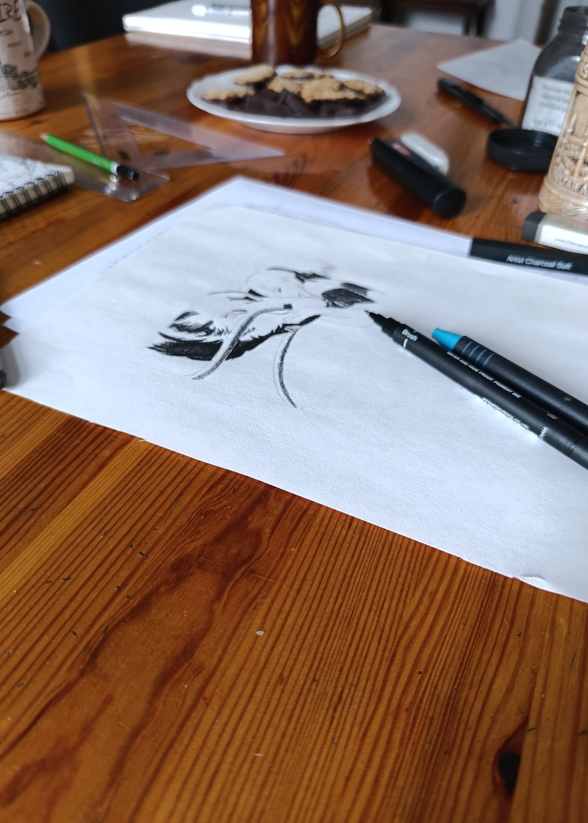 A black ink sketch of a dog on a sheet of white paper resting on a wooden table, with pens and other desk items in the background.