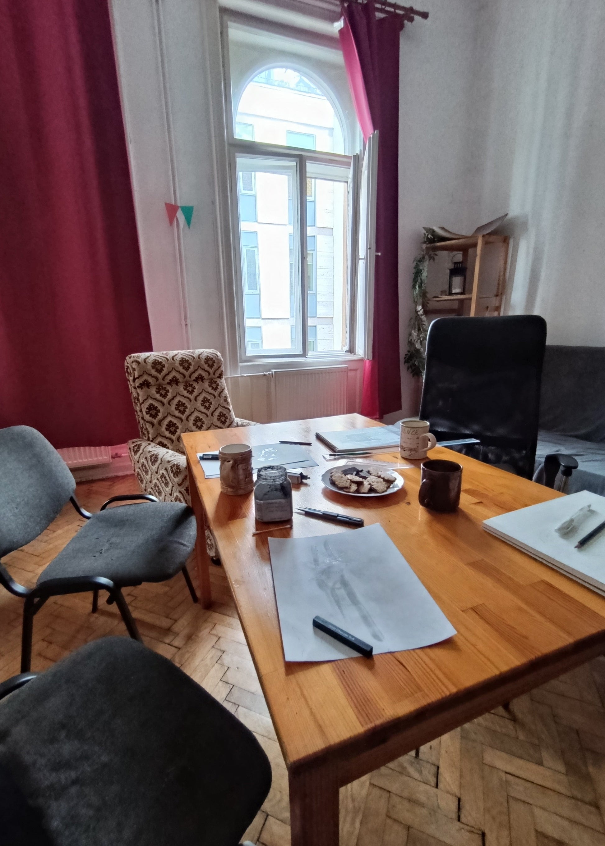 A wooden writing desk in a room with patterned and black office chairs, a window with red curtains, and papers on the tabletop.