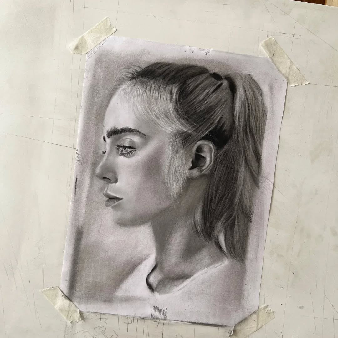 A detailed charcoal portrait drawing of a woman with her hair in a ponytail, taped onto a larger sketchbook with tape at the corners.