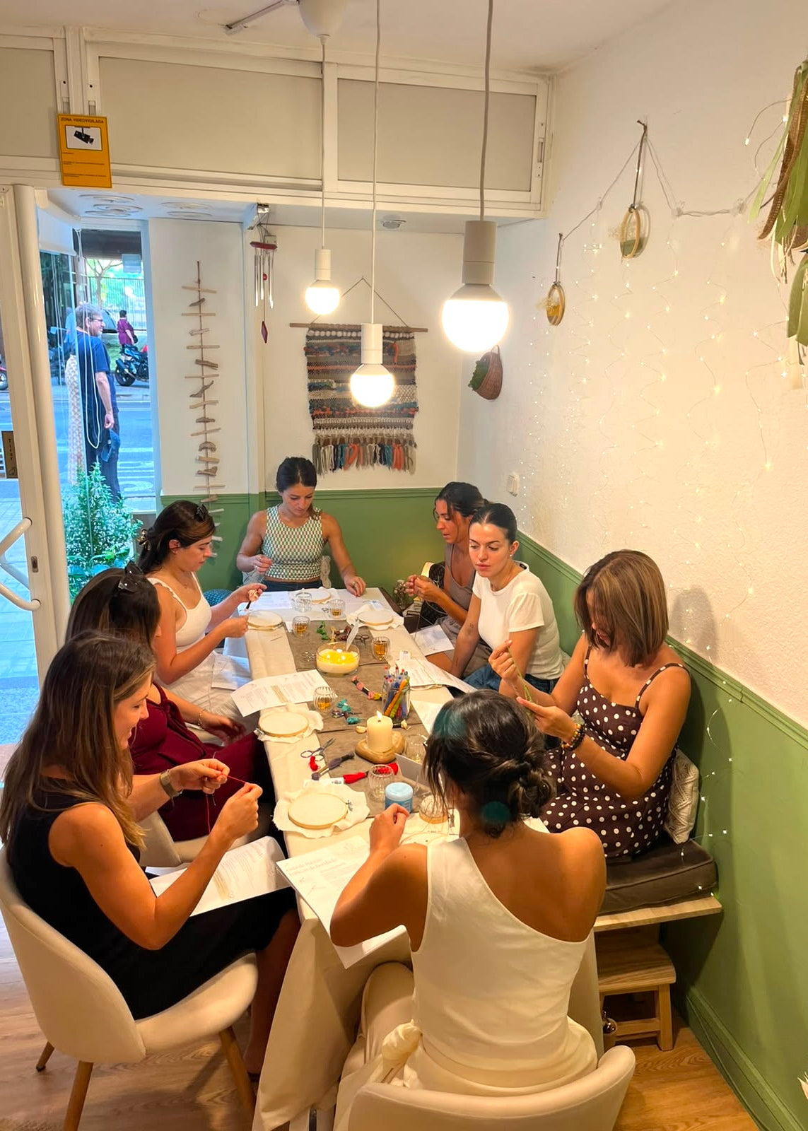 A group of women gathered around a table in a cozy, brightly lit cafe or workshop space with green and white walls, pendant lights, and string lights.