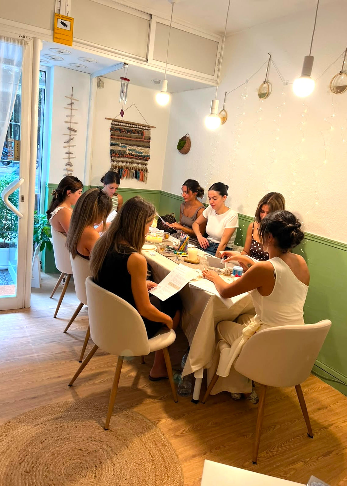 A group of women seated around a table in a bright, modern room with green and white walls, wooden floors, pendant lights, and string lights. They appear to be engaged in a workshop, meeting, or class activity, with papers and materials on the table.