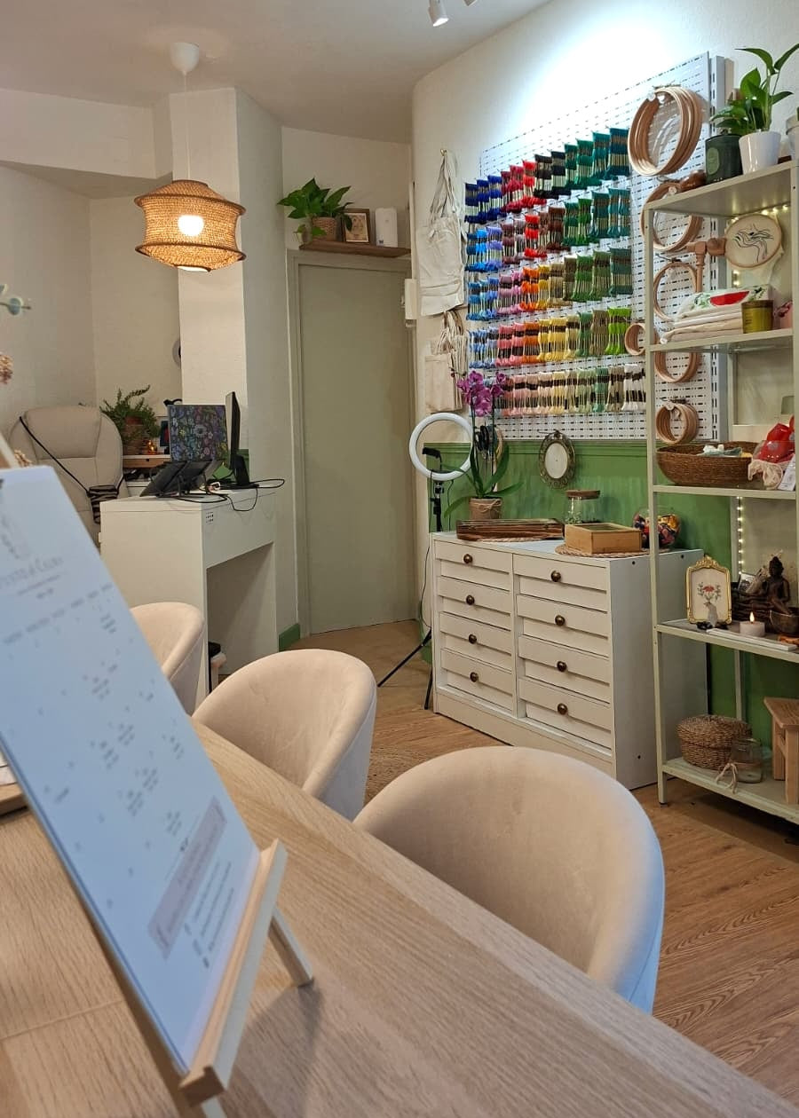 A bright, modern craft studio or sewing room with a large wall-mounted pegboard display of colorful thread spools. The room features white furniture, light wood floors, a wooden table in the foreground with a whiteboard on an easel, and a green accent wall.