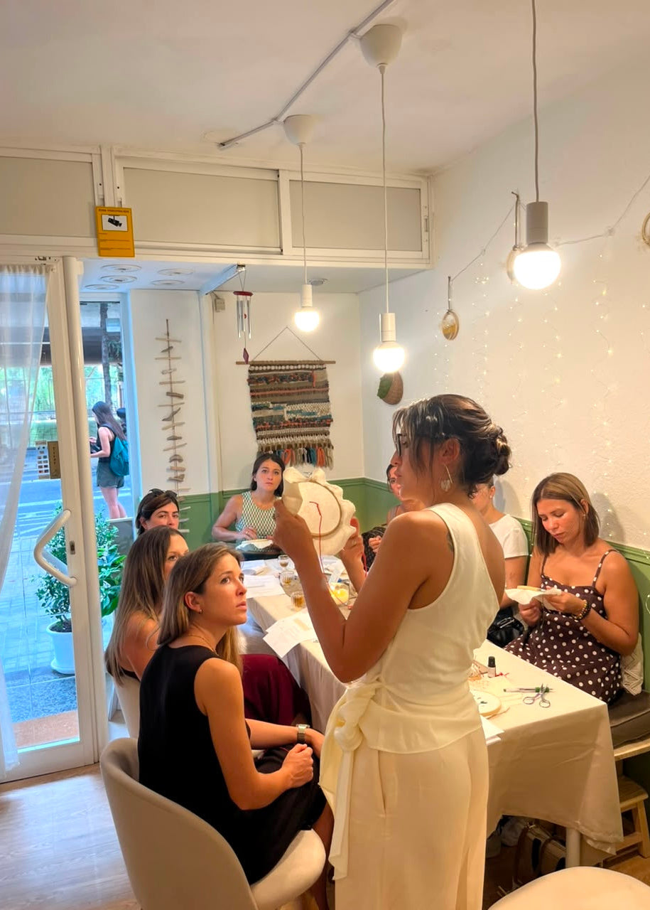 A woman in a white sleeveless dress holds a light-colored hat while standing in a bright, modern dining or workshop space with several seated women around her.