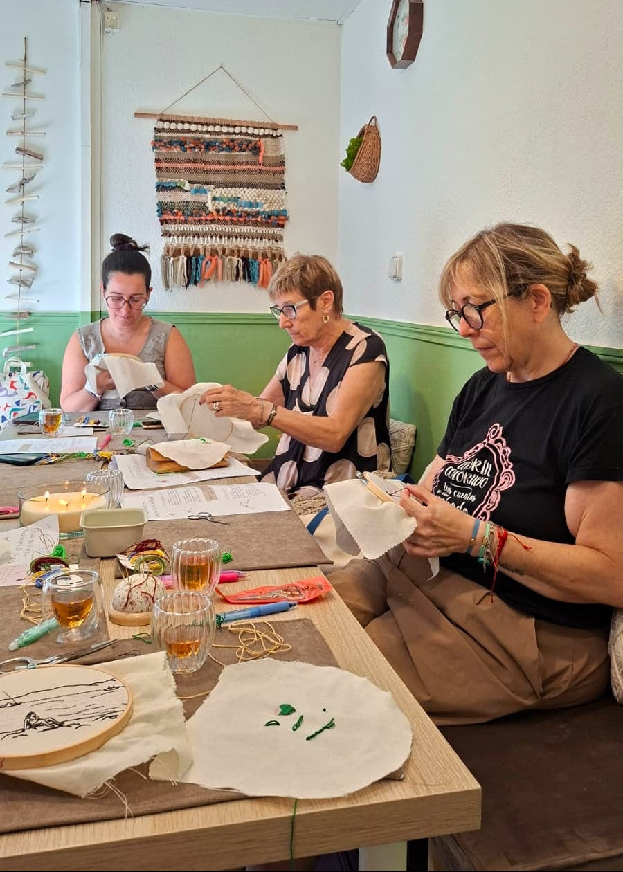 Three women seated around a table in a bright room, engaged in embroidery or textile crafting with papers, threads, and small supplies spread across the table.