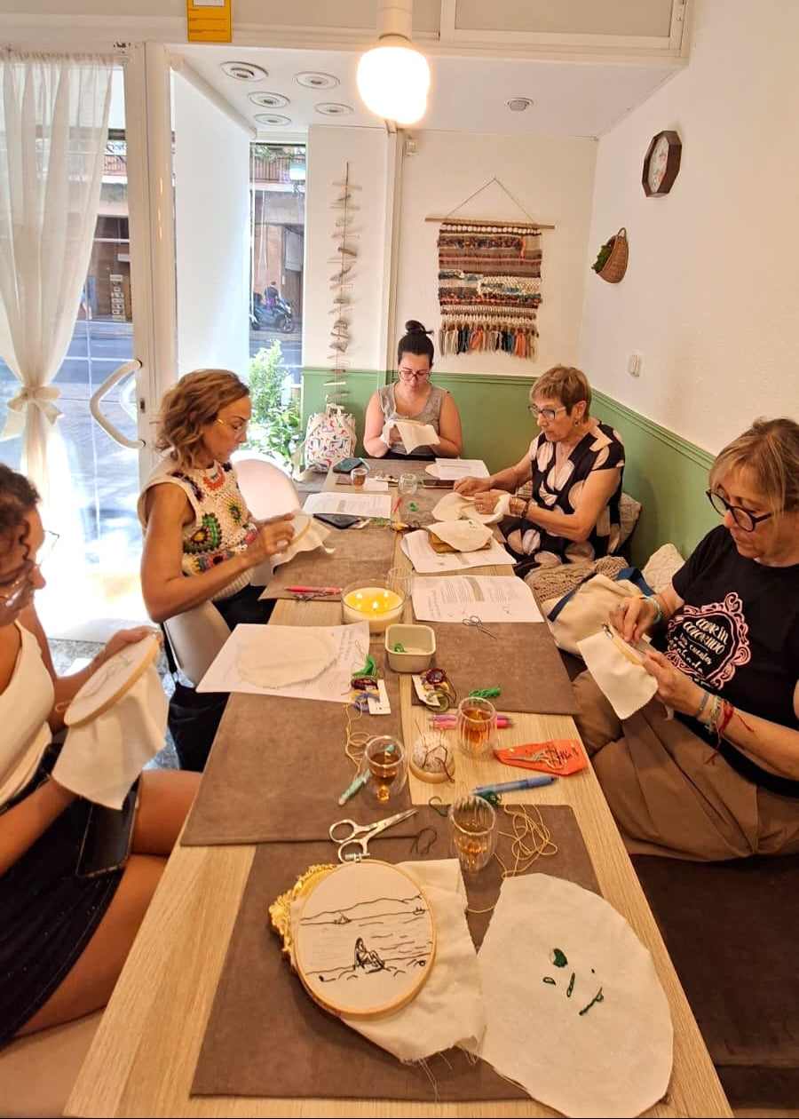 A group of five women sitting around a wooden table in a bright, cozy room, engaged in embroidery or sewing activities with threads, hoops, and papers. The room features white walls, a green lower panel, a hanging light bulb, and a window with sheer curtains.
