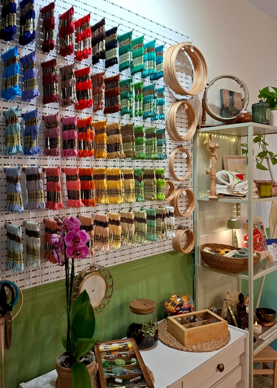 A styled interior display featuring a large wall-mounted pegboard filled with many colorful hanging items, likely craft or accessory packs. To the right, a white shelving unit holds mirrors, books, and decorative objects.