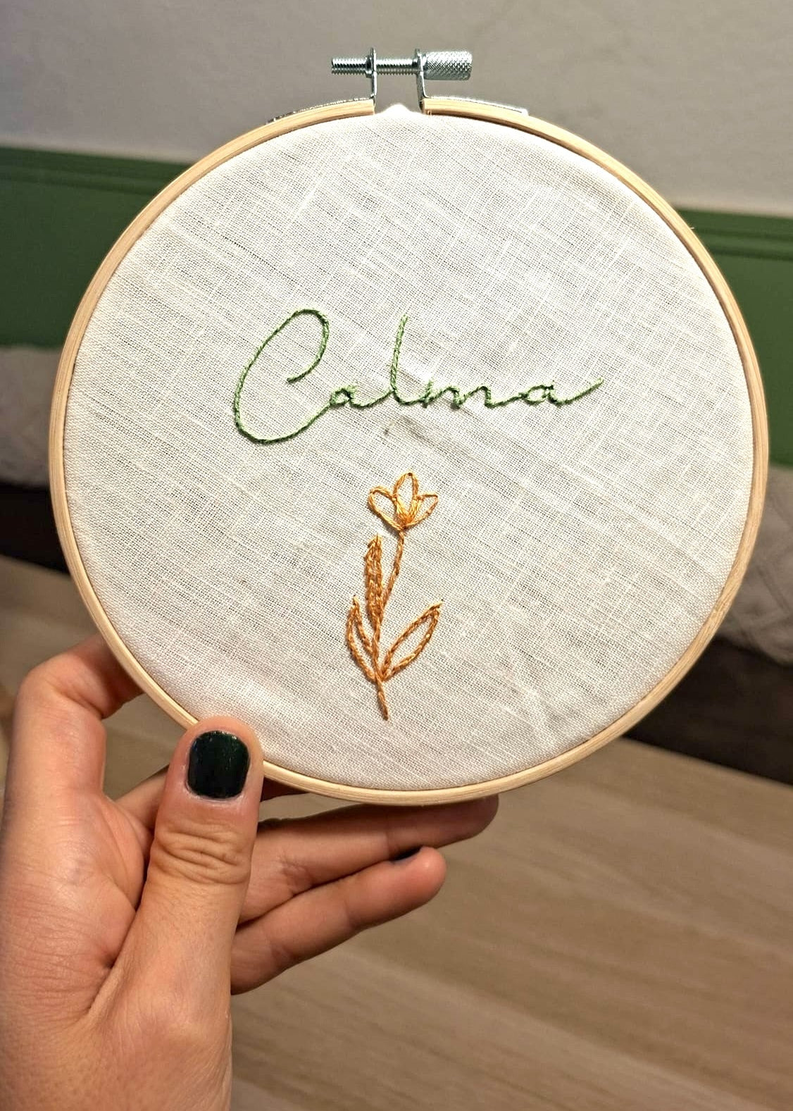 A hand with black nail polish holds a wooden embroidery hoop containing light beige fabric with green and gold embroidered text reading 'Calma' and a small flower.