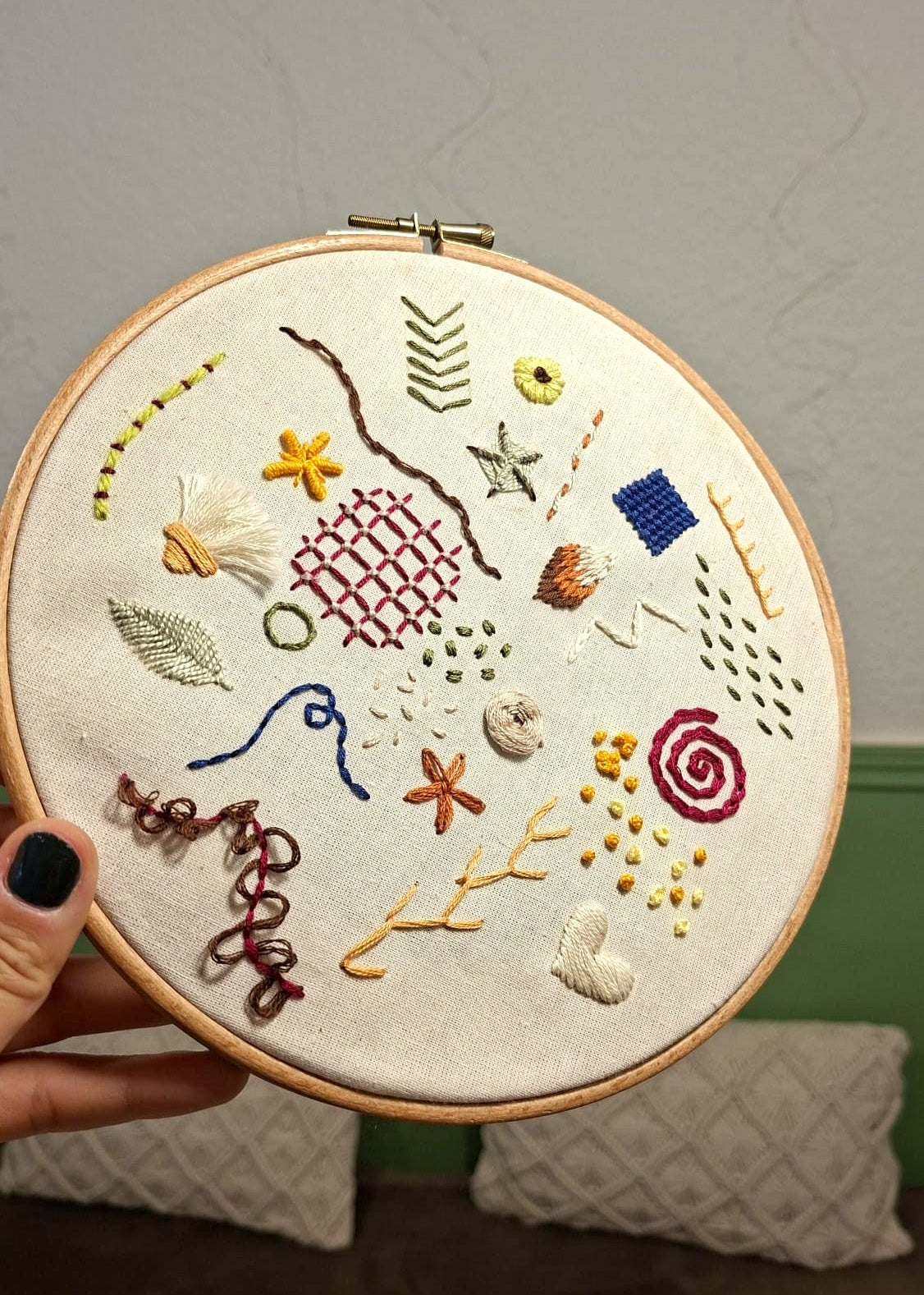 A hand with black nail polish holds a wooden embroidery hoop containing a colorful assortment of embroidered motifs, including flowers, leaves, geometric patterns, and abstract designs on cream fabric, set against an indoor background.