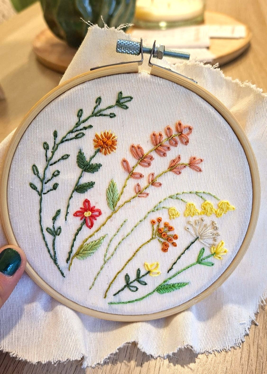 A round embroidery hoop holding white fabric with colorful floral and leaf stitching, photographed on a wooden table with a hand holding the hoop and blurred home decor in the background.