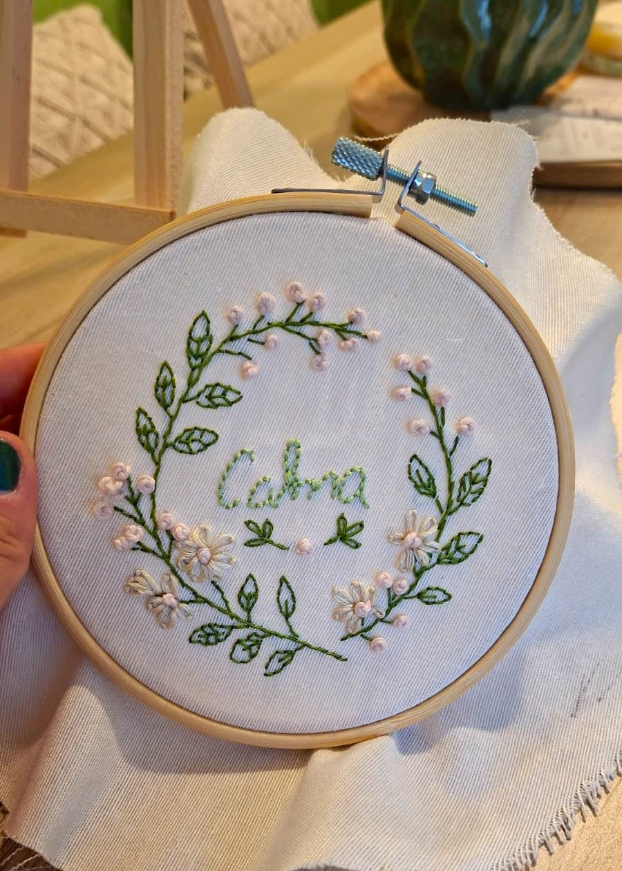 A close-up of a hand holding a wooden embroidery hoop with a finished floral wreath embroidery design on white fabric. The design features green leaves, small pink flowers, and the name 'Caba' stitched in green in the center.
