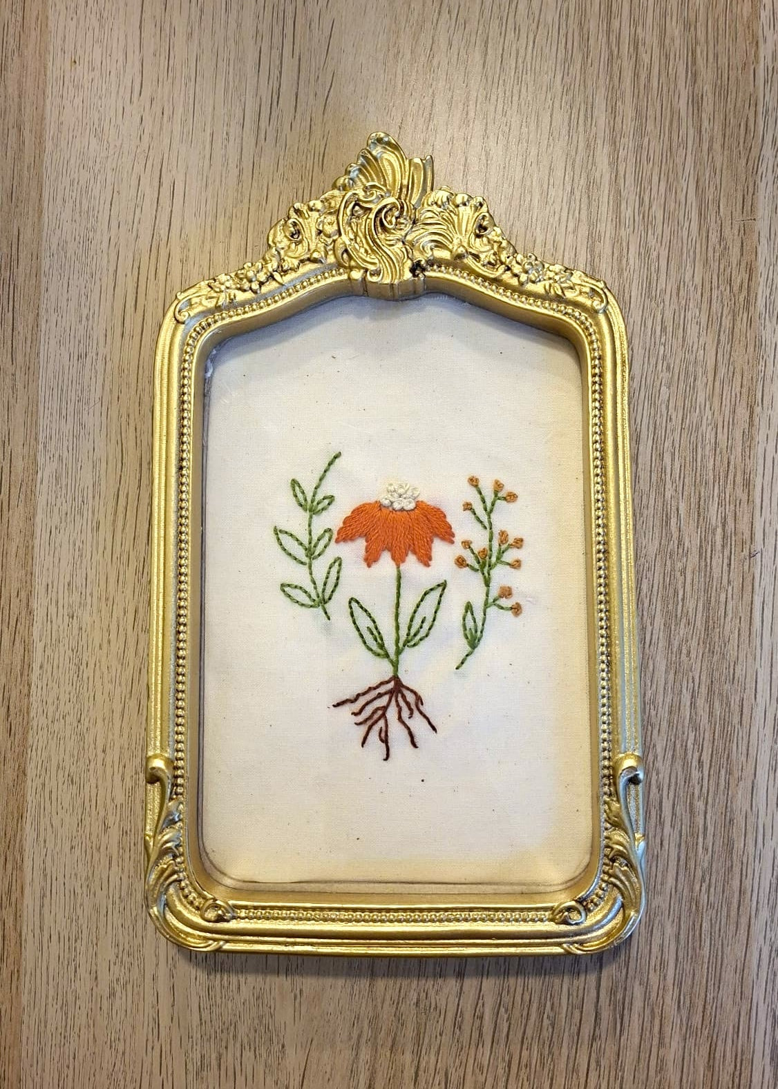 An ornate gold picture frame with a white linen insert featuring a hand-embroidered orange flower with green leaves and brown roots, displayed on a light wood background.