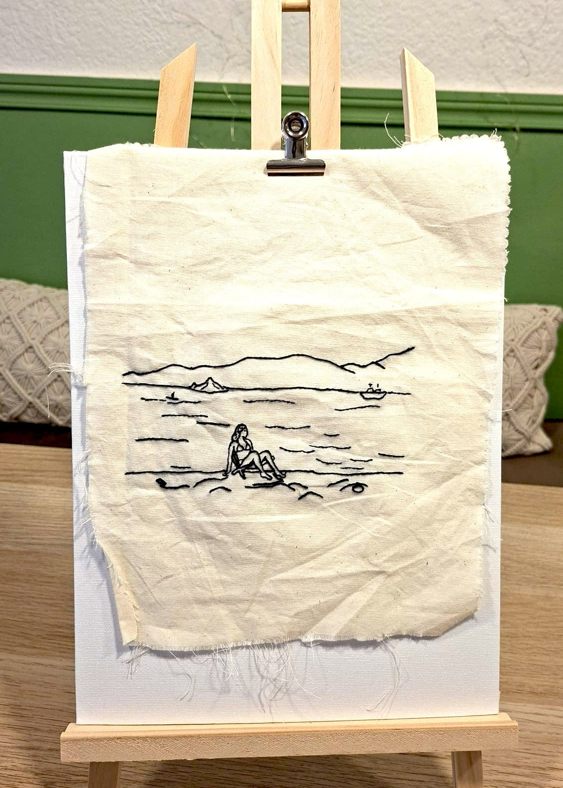 A square piece of off-white fabric with a simple black line drawing of a seated figure by water, displayed on a small wooden easel against a green wall and wood floor.
