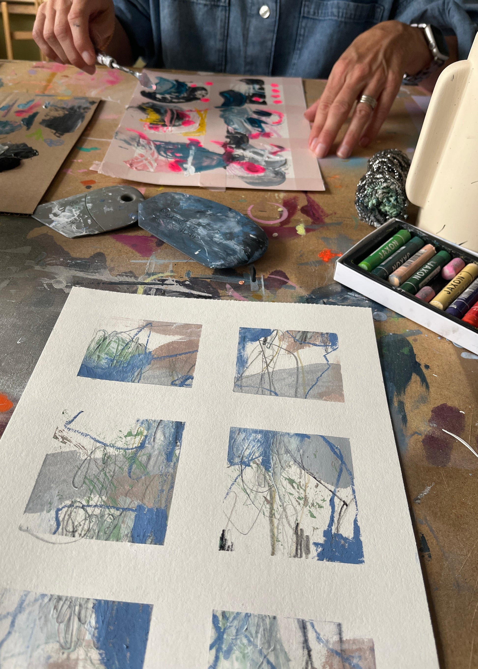 A person's hands and torso are visible at a painted table, working on abstract art pieces. In the foreground, a sheet of textured paper holds several painted square sections with blue, gray, beige, and green brushstrokes.