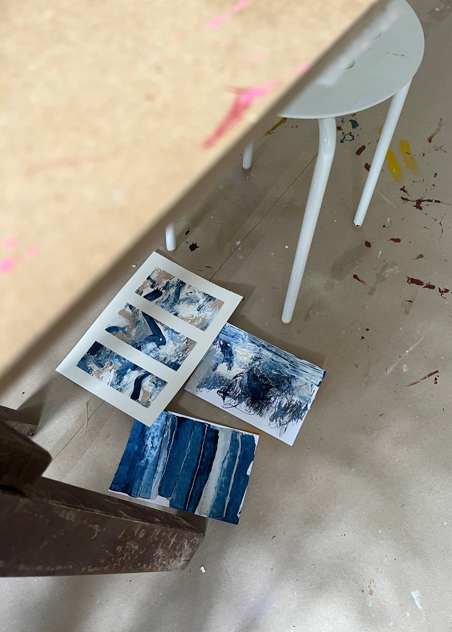Three abstract blue and white painted art prints or cards scattered on a paint-splattered floor beneath a table and stool in an artist's studio.