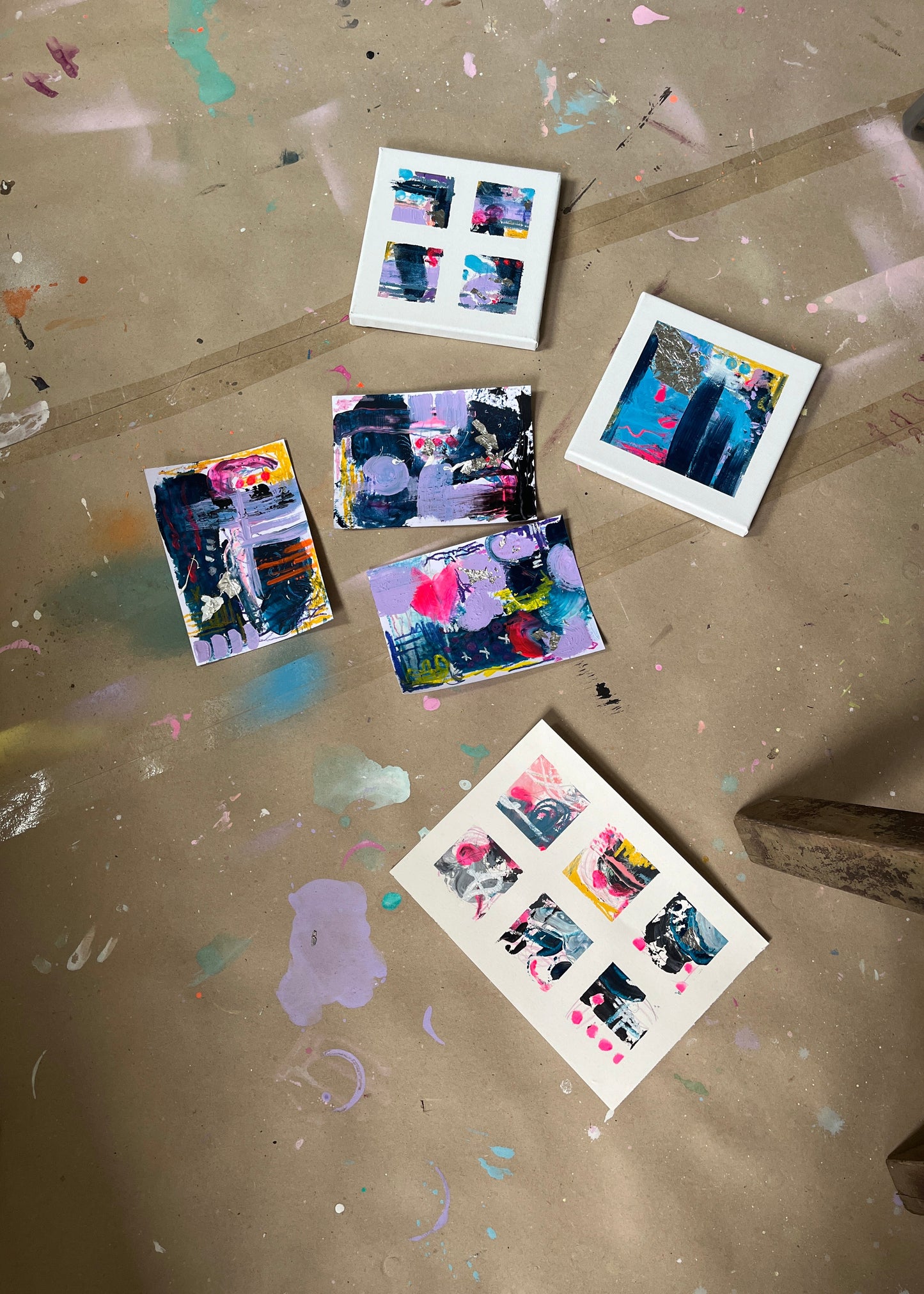 Five small abstract paintings in white frames and one larger sheet of six paintings, arranged on a paint-splattered brown floor.
