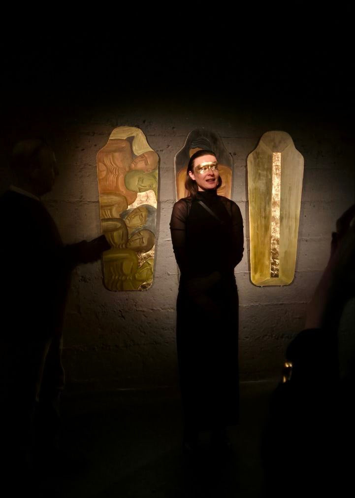 A person in a black outfit stands in a dimly lit gallery or exhibition space, facing three illuminated decorative panels mounted on a wall. The scene is very dark, with the central figure and parts of the surroundings barely visible, creating a moody, low-key atmosphere.