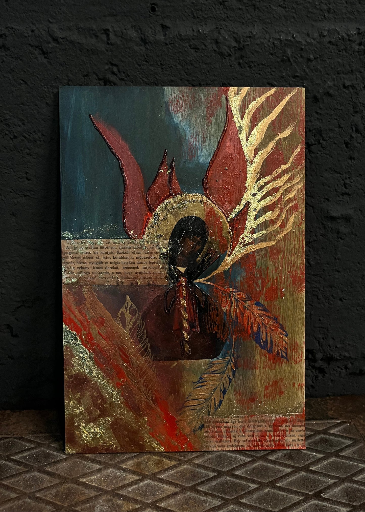 An abstract mixed-media painting of an angel with red wings and a halo, displayed on a textured dark wall.