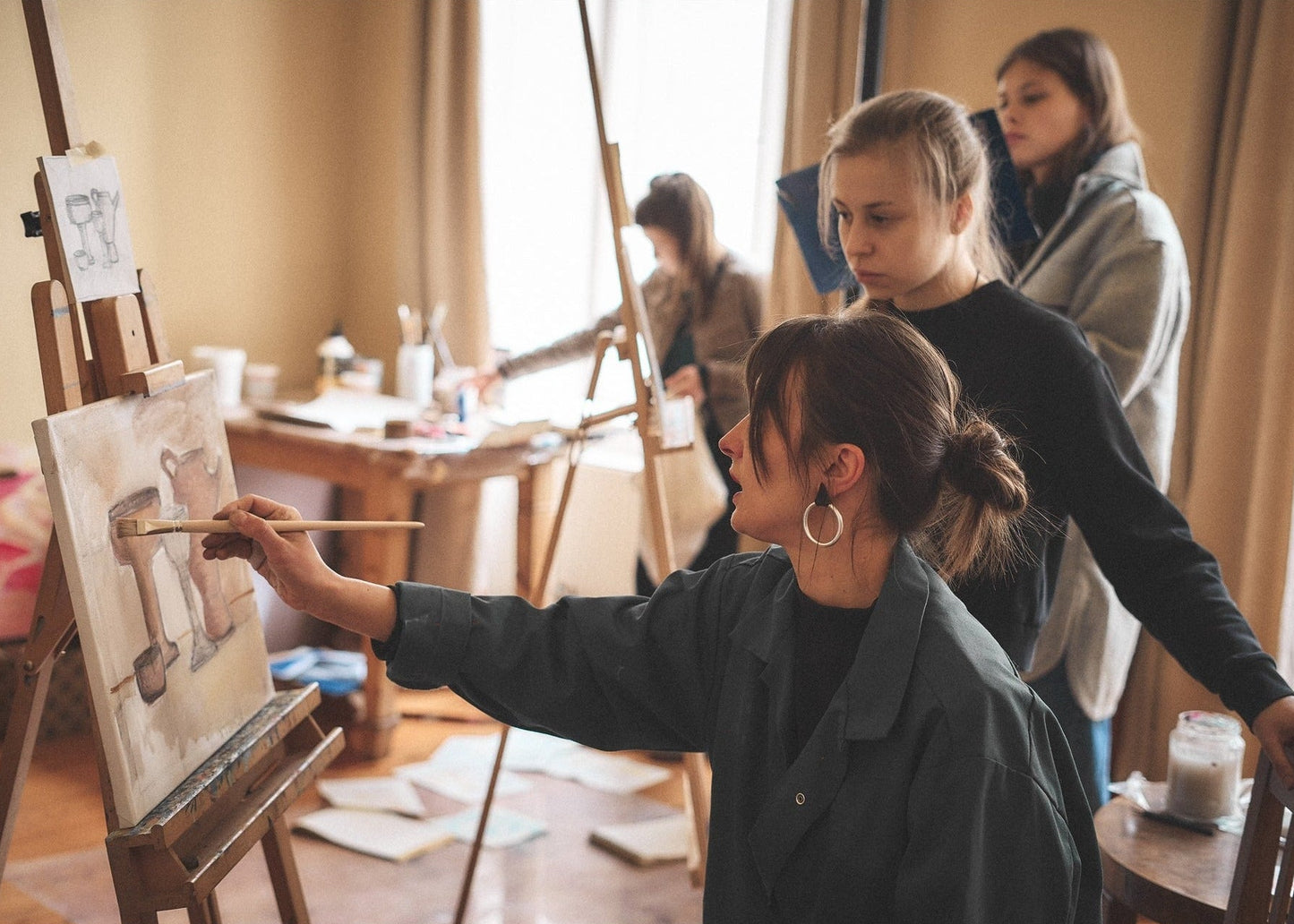 A woman in a dark green jacket paints on an easel in a warm, sunlit art studio, with two other women standing nearby and another artist visible in the background.