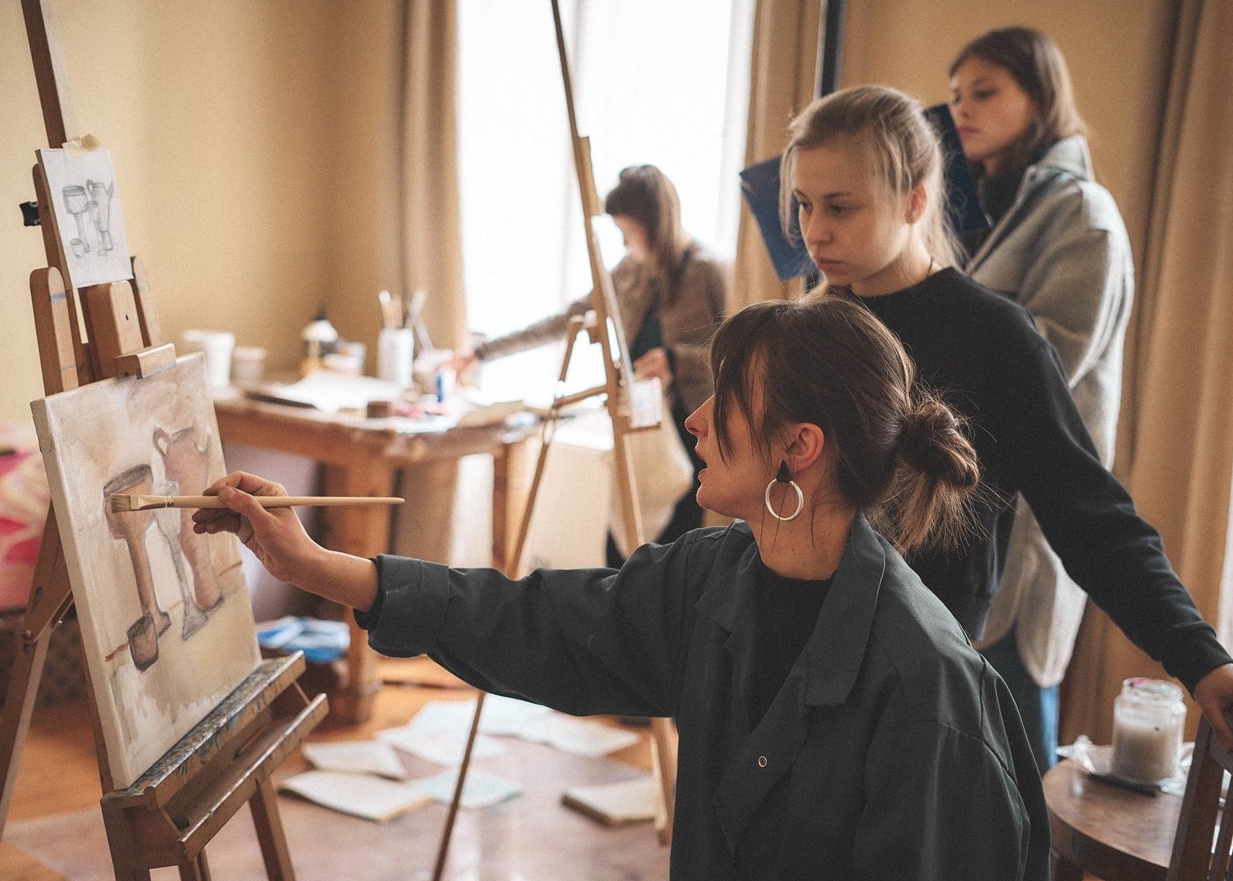 A woman in a dark green jacket paints on an easel in a warm, sunlit art studio, with two other women standing nearby and another artist visible in the background.