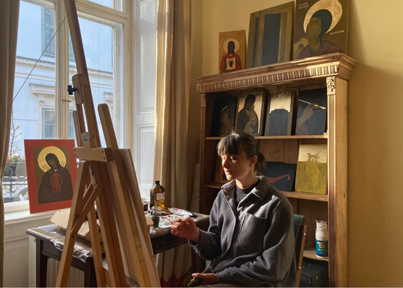 A woman sits at a table in a sunlit artist studio, painting at an easel. A wooden bookshelf behind her holds several framed religious icons and paintings.