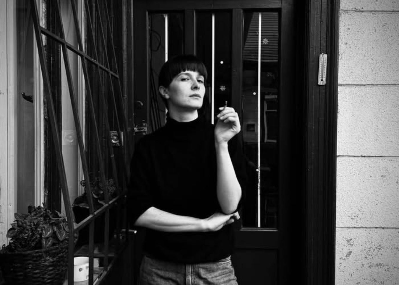 A black and white portrait of a person with short dark hair wearing a black turtleneck sweater and light-colored jeans, standing in front of a dark doorway.