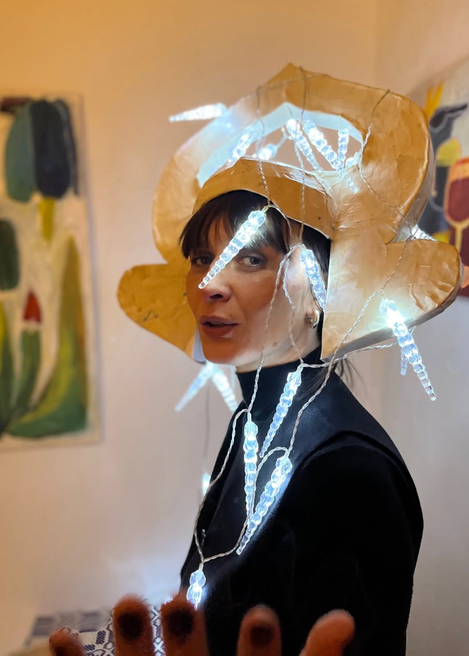 A person wearing a large sculptural hat made of gold-toned material with hanging white LED light tubes, photographed indoors against a beige wall with framed artwork in the background.