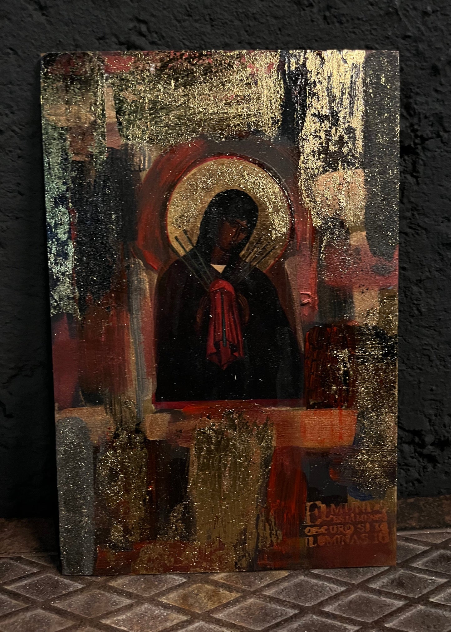 An abstract religious icon painting of a robed figure with a halo, rendered in dark red, black, gold, and brown tones, displayed against a dark textured wall.