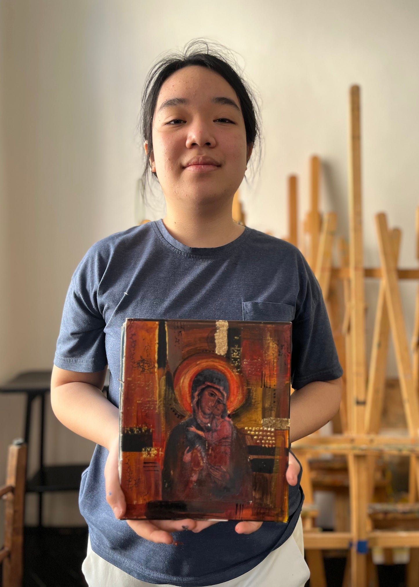 A young woman in a blue t-shirt holds up a small, colorful religious painting of the Virgin Mary with a golden halo in an art studio setting.
