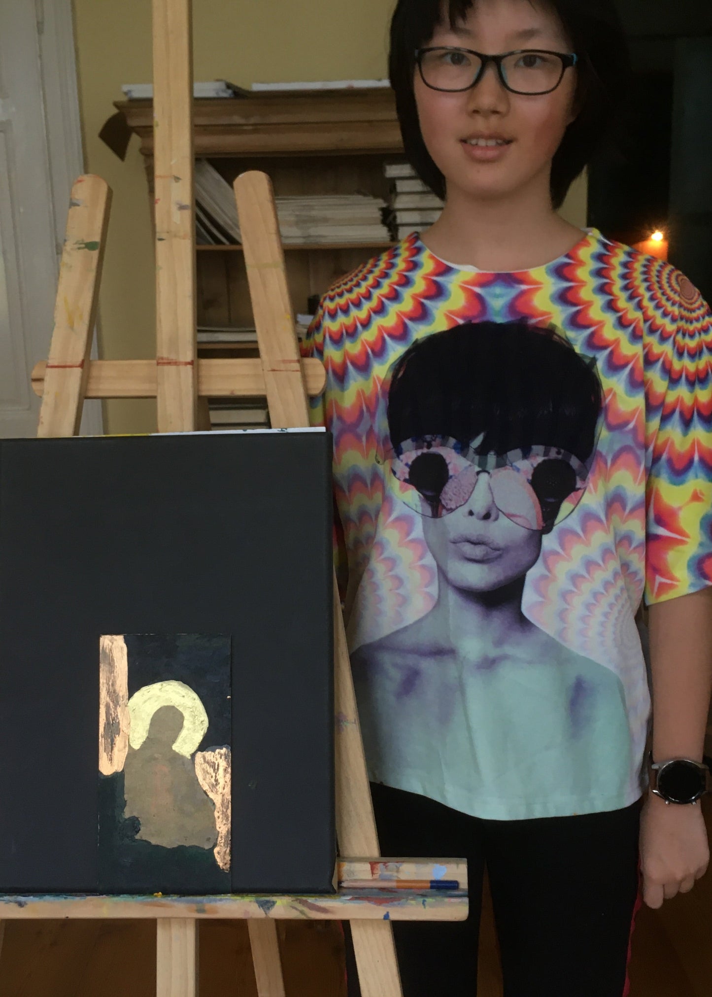 A person wearing a colorful rainbow spiral-patterned t-shirt with a graphic of a woman in sunglasses, standing next to a black canvas painting on a wooden easel in an indoor room.