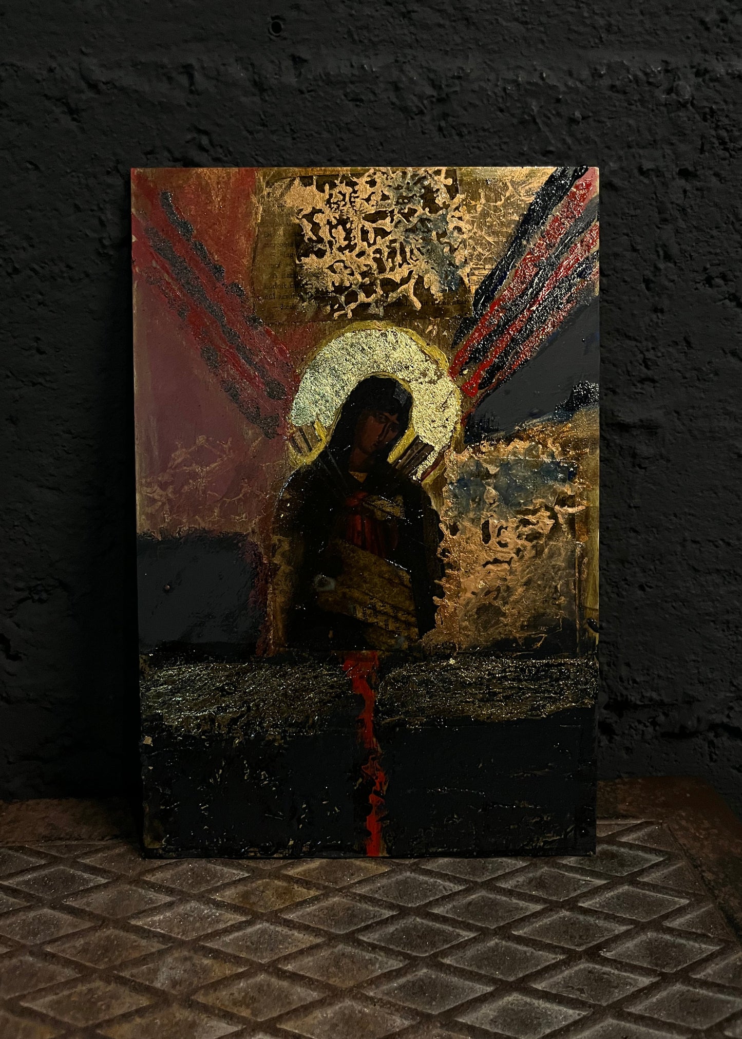 An abstract religious canvas painting leaning against a dark textured wall on a tiled floor. The artwork features a haloed figure in black and gold, with red and black brushstrokes radiating outward.