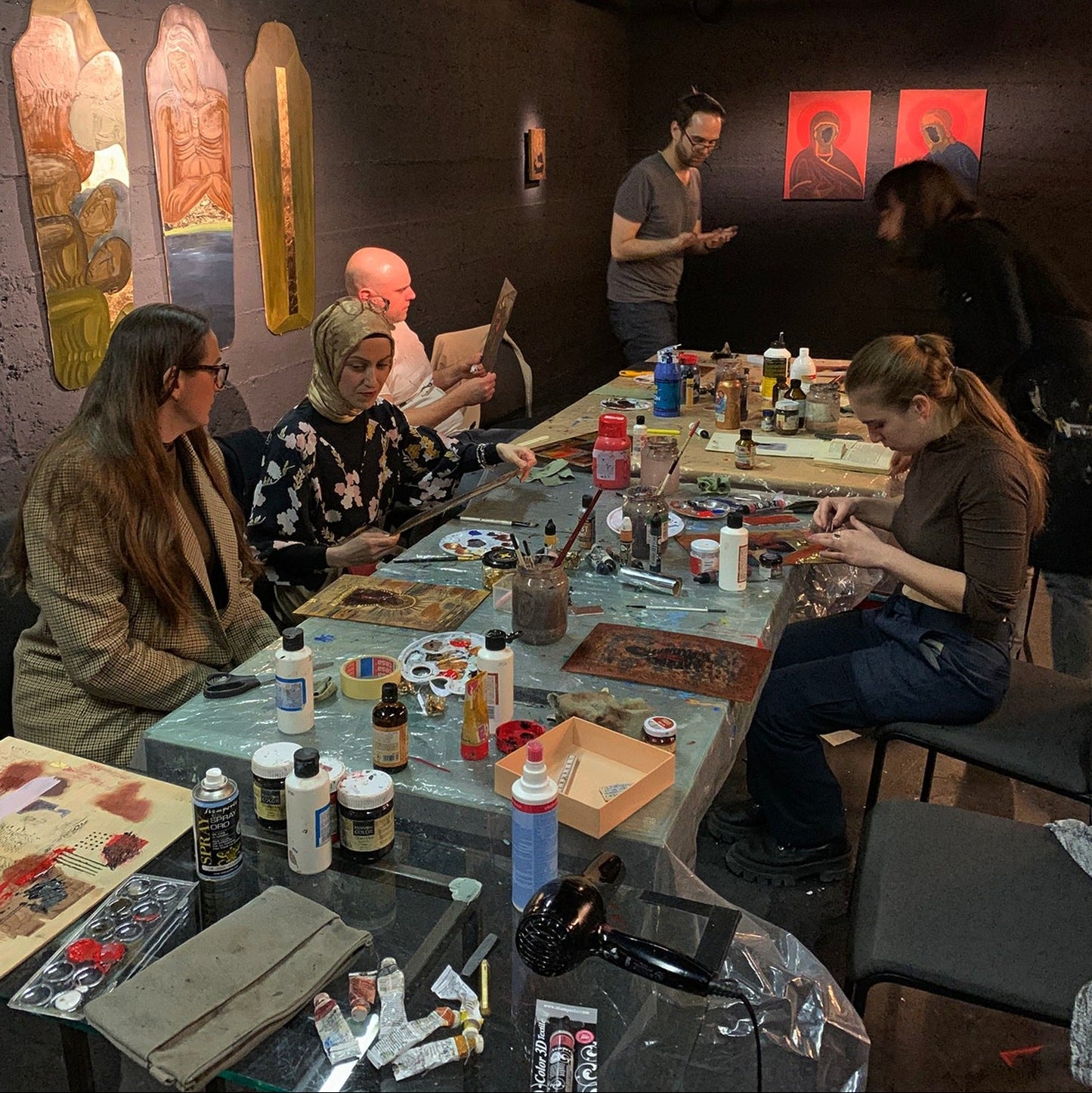 A group of people gathered around a long table in a studio or workshop, working on paintings and handling art supplies. The room has dark walls with several colorful framed artworks hanging on them, and the table is covered with paint cans, brushes, palettes, and other painting materials.