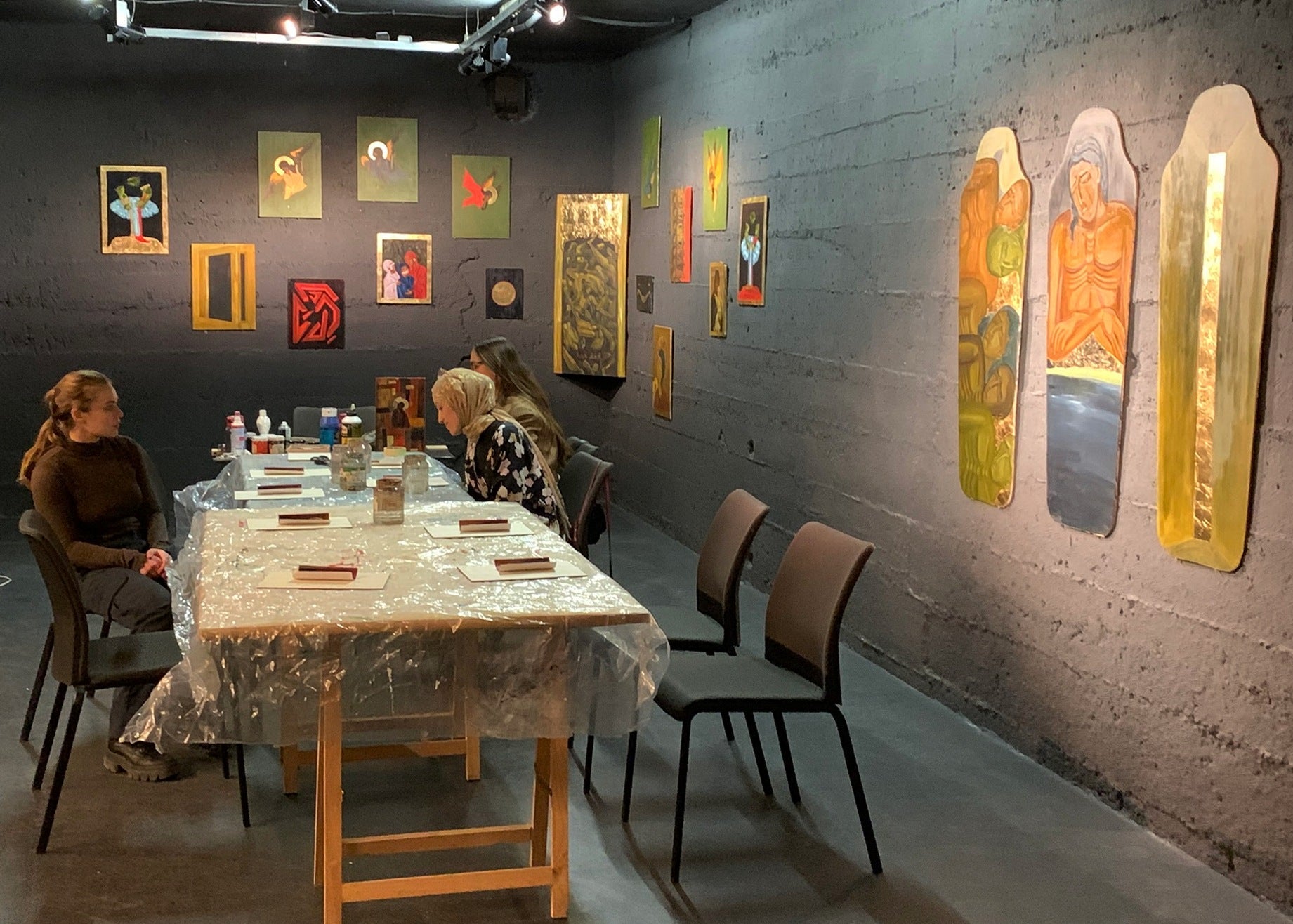 An indoor art gallery or studio space with a long table covered in a clear plastic sheet, three seated people at the table, and multiple colorful framed artworks hanging on dark gray walls.