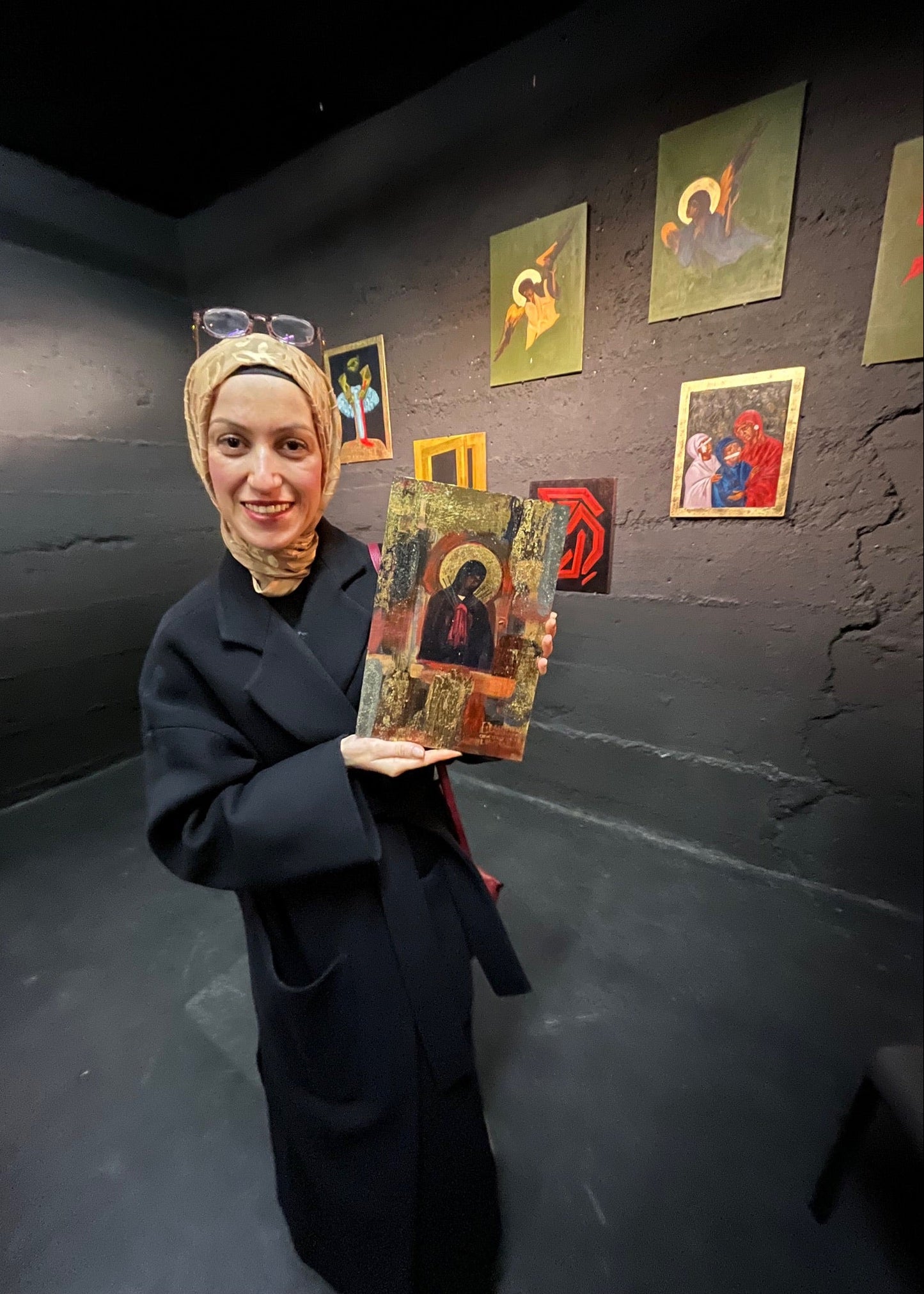 A woman wearing a beige headscarf and a long black coat holds up a traditional religious icon painting in a gallery with dark walls and other framed art in the background.