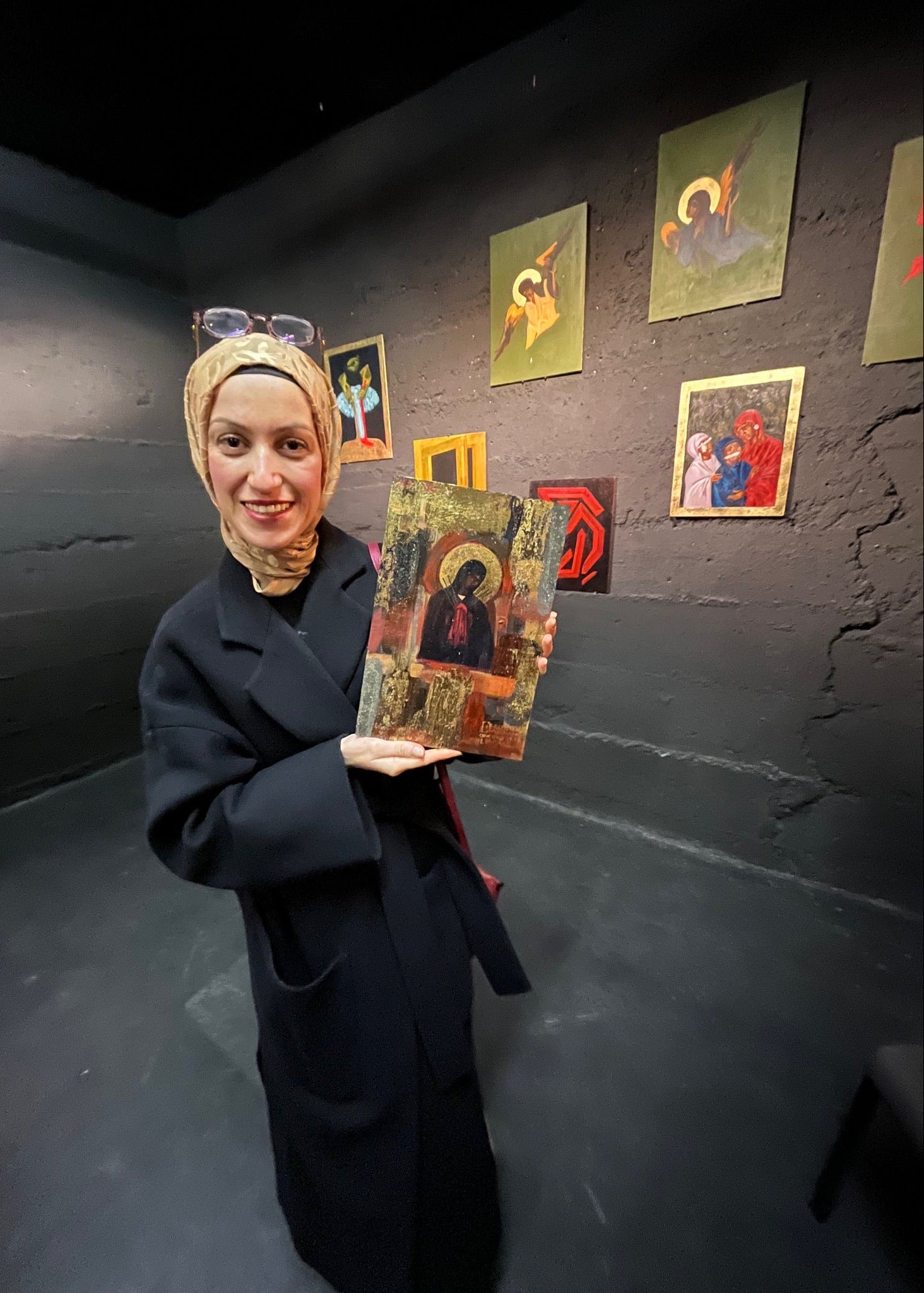 A woman wearing a beige headscarf and a long black coat holds up a traditional religious icon painting in a gallery with dark walls and other framed art in the background.