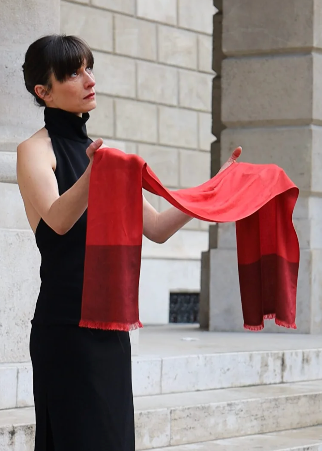 A woman in a black sleeveless dress holds up a red scarf with a solid red top half and a darker red bottom half, standing in front of a light stone building.