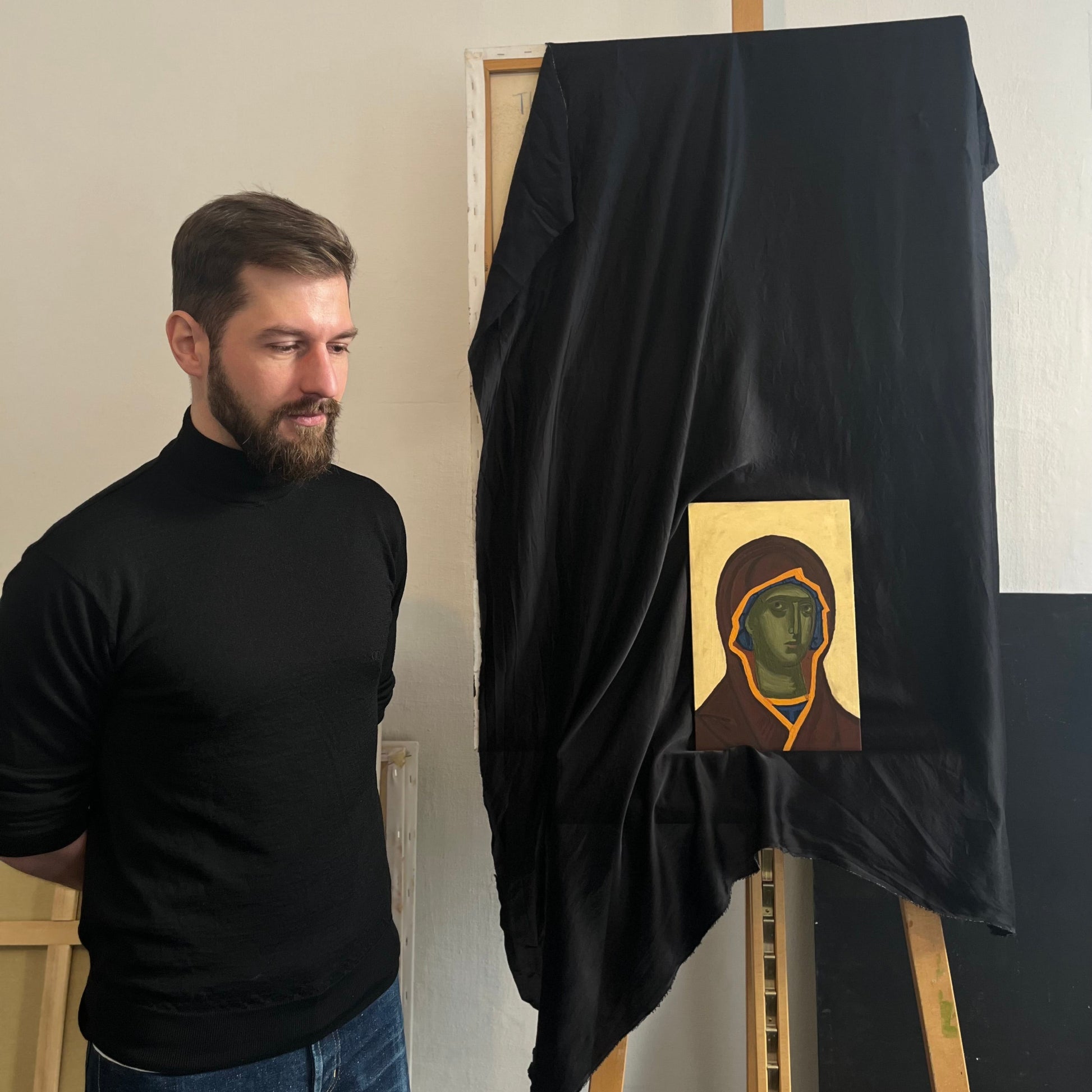 A man in a black turtleneck and blue jeans stands beside an easel draped in black fabric, revealing a small religious icon painting with a gold background and a robed figure.