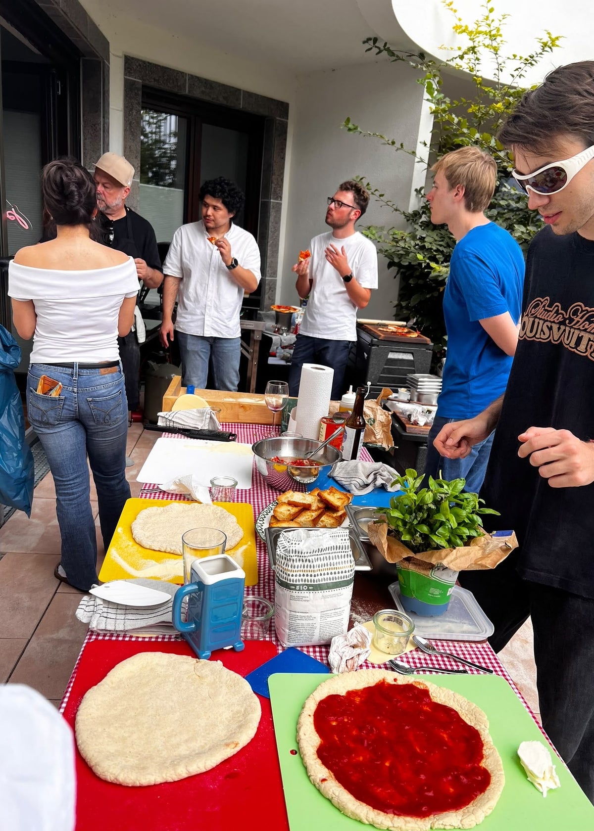 A group of young men gathered around a table outdoors, preparing pizza dough and spreading tomato sauce on flatbreads. The table is covered with pizza ingredients and utensils, set against a patio background with people interacting.