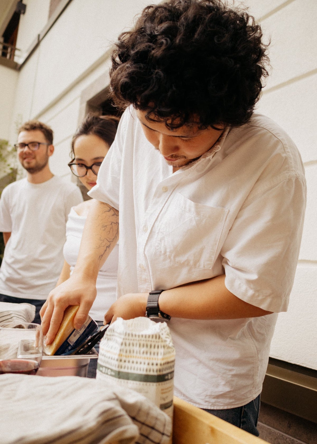 A person in a white short-sleeve button-up shirt is leaning over a table, holding a sponge and interacting with items on a counter. Two other people in white shirts are visible in the background against a light-colored exterior wall.