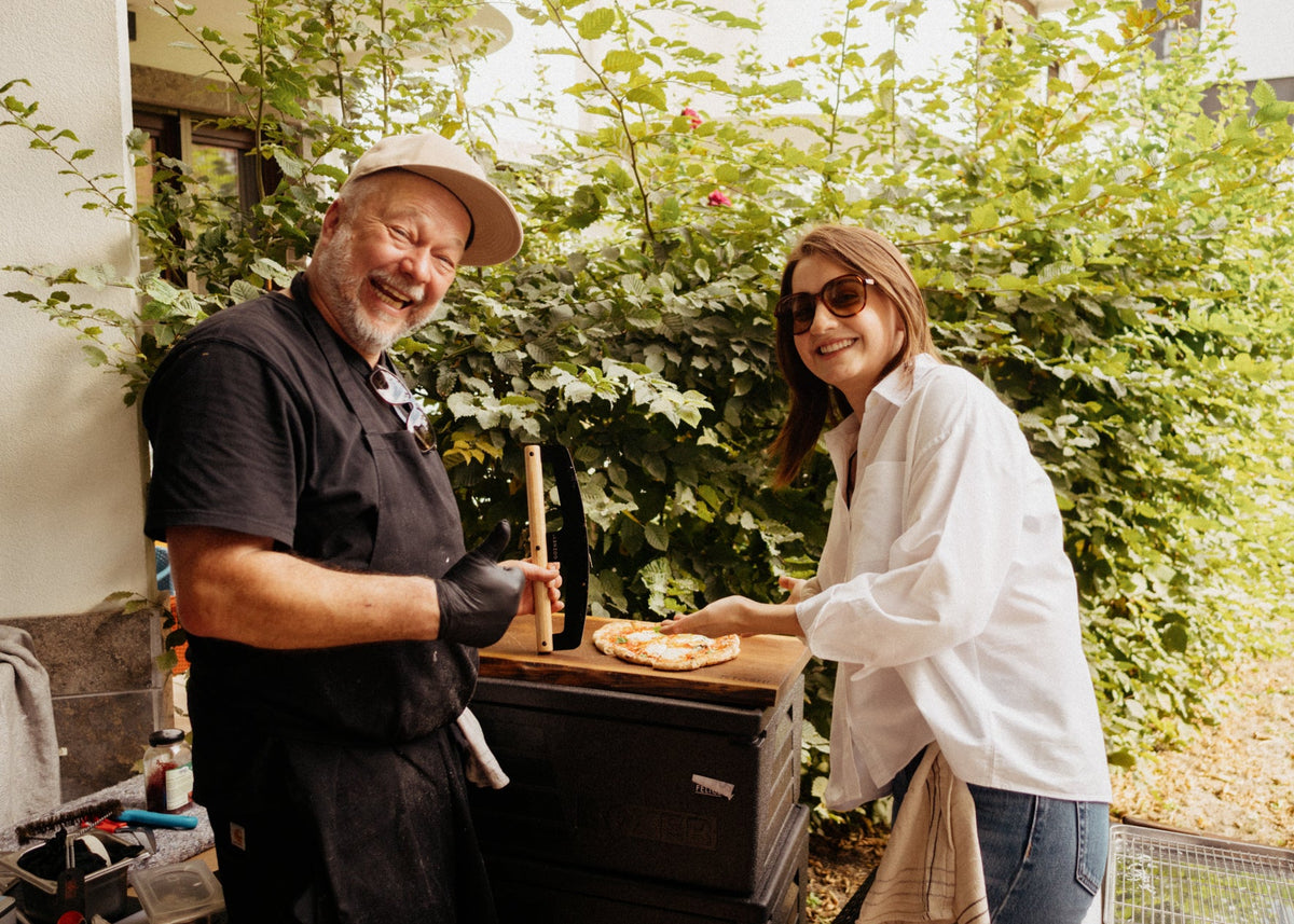 A man in a black chef's coat and white cap smiles while holding a pizza peel with a pizza on it, next to a woman in a white shirt and sunglasses who is handing him a slice of pizza. They are outdoors in front of a lush green garden wall.