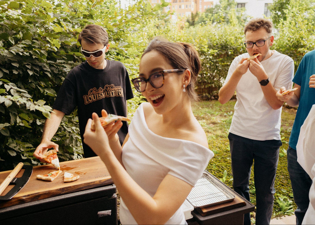 A woman in a white off-the-shoulder top and glasses laughs while holding a slice of pizza outdoors at a barbecue grill, with three men eating pizza in the background among green bushes.