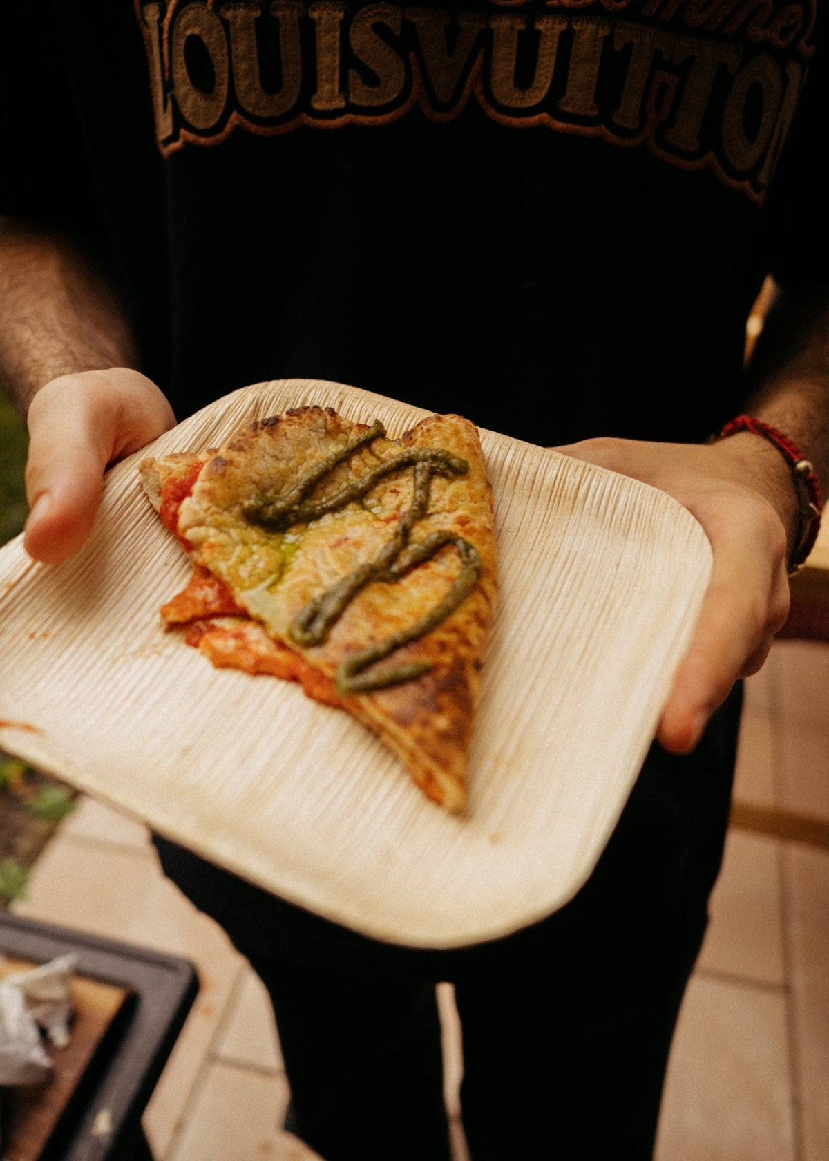 A person holding a triangular paper plate with a slice of pizza topped with green sauce, wearing a black Louis Vuitton t-shirt.