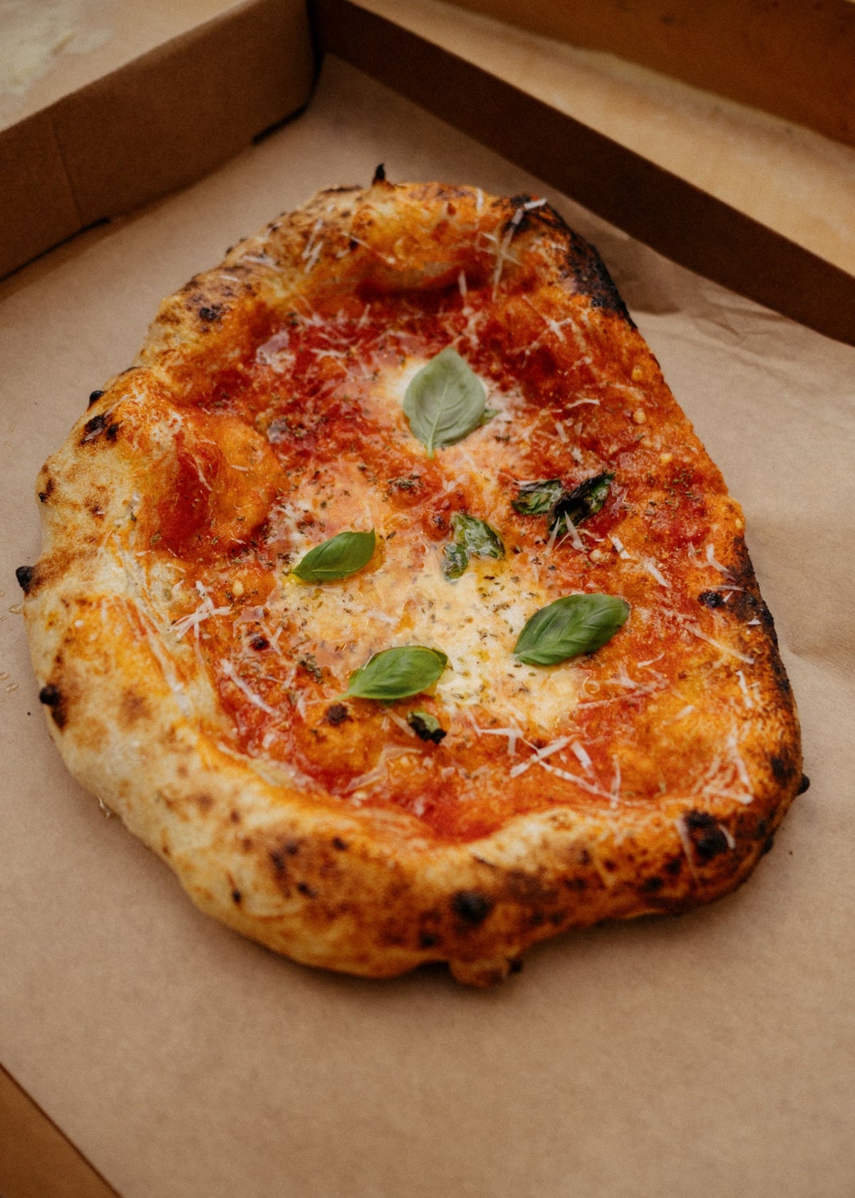 A freshly baked artisanal pizza with a charred crust, topped with tomato sauce, melted cheese, and fresh basil leaves, presented in a cardboard pizza box on parchment paper.