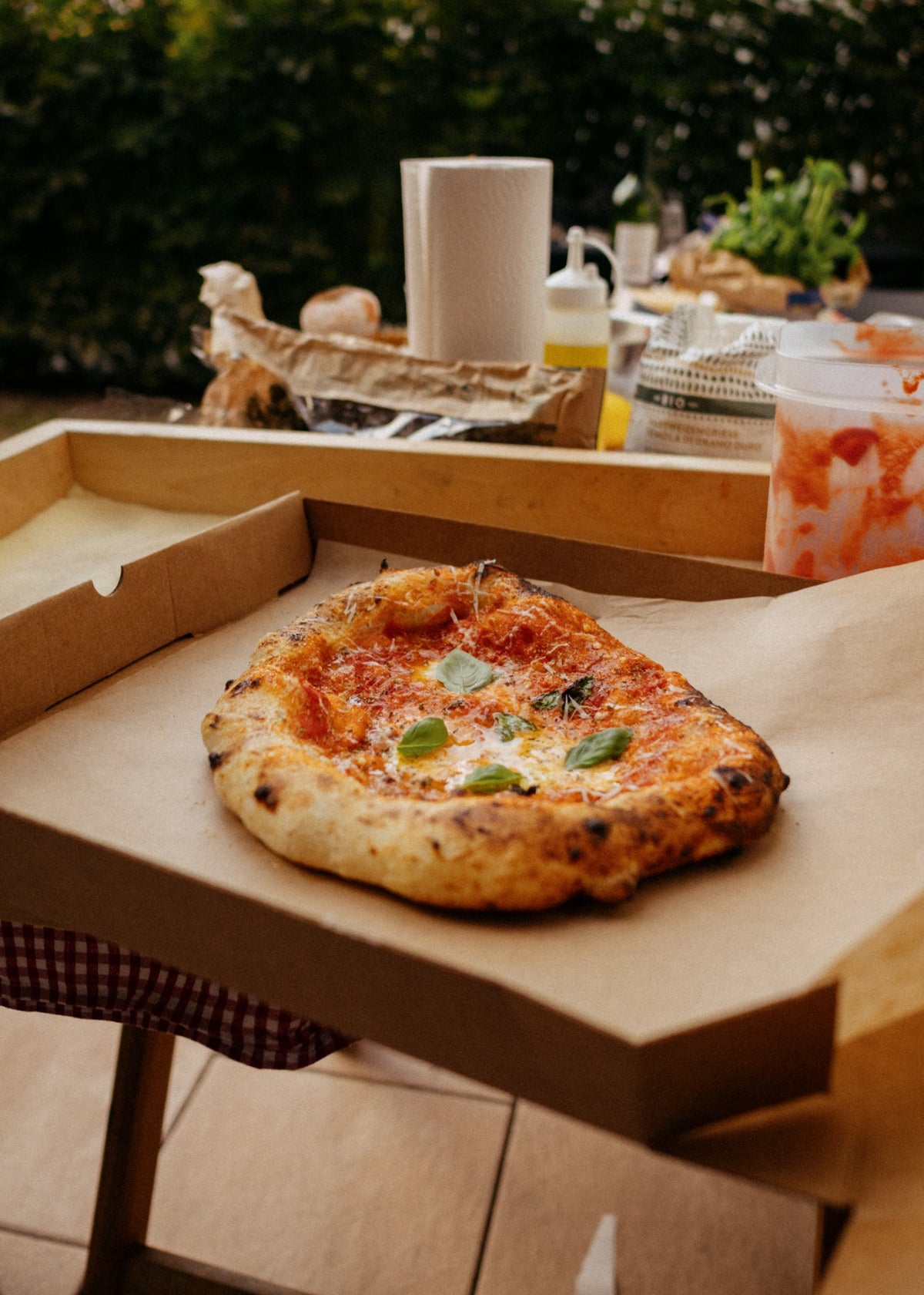 A freshly baked pizza with basil on a wooden board inside an open cardboard pizza box, set on an outdoor patio table with dining items in the background.