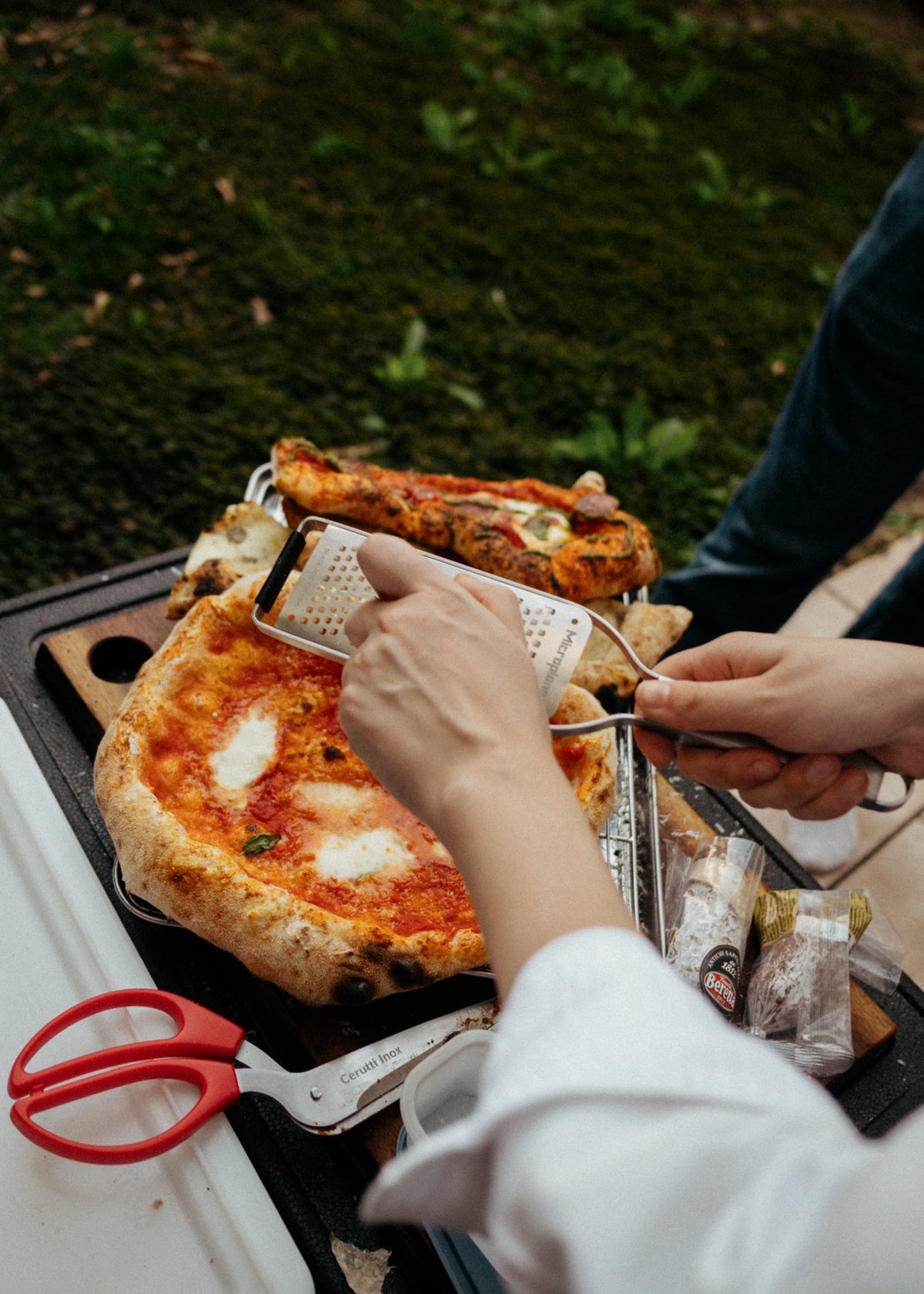 A close-up lifestyle shot of a person grating cheese onto a freshly baked pizza outdoors. The scene includes another pizza, kitchen shears, and a person's hands and legs in a real outdoor environment.