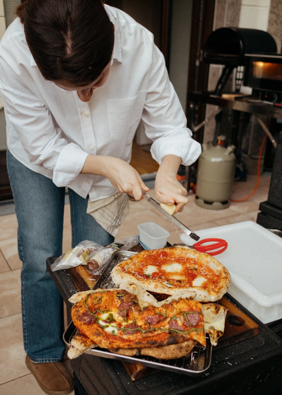 A person in a white button-up shirt and blue jeans slices a large freshly baked pizza on a metal tray at an outdoor cooking station.