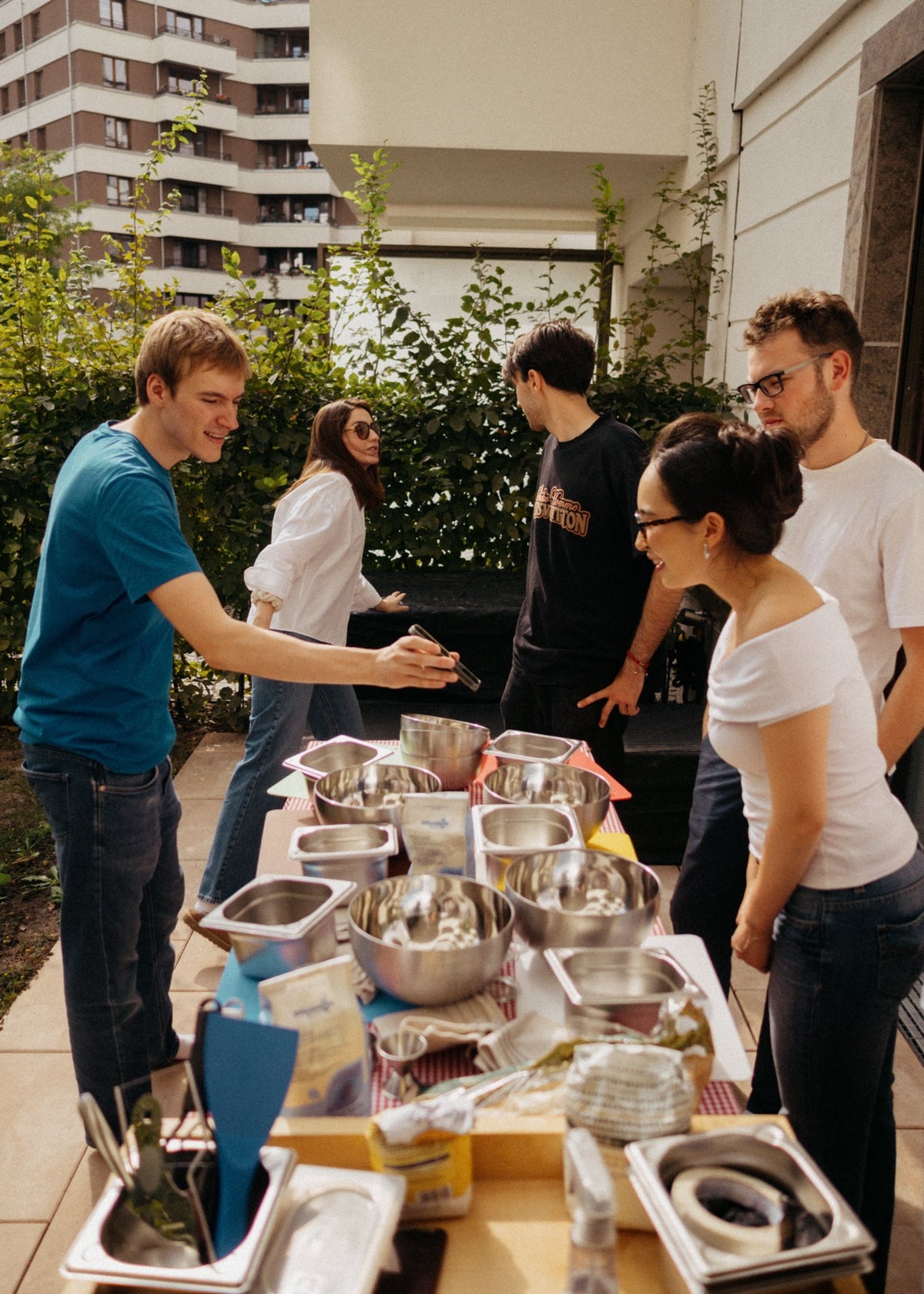 A group of five young people gathered outdoors on a patio around a table filled with stainless steel mixing bowls, utensils, and baking ingredients. Two individuals are actively mixing ingredients in large metal bowls, while the others observe and assist.
