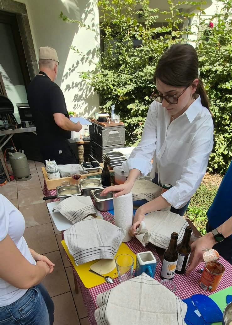 A group of people gathered around a table outdoors, handling stacks of round white woven coasters or mats on a red-and-white checkered tablecloth. Bottles, a cup, and other items are visible on the table, with patio furniture and greenery in the background.