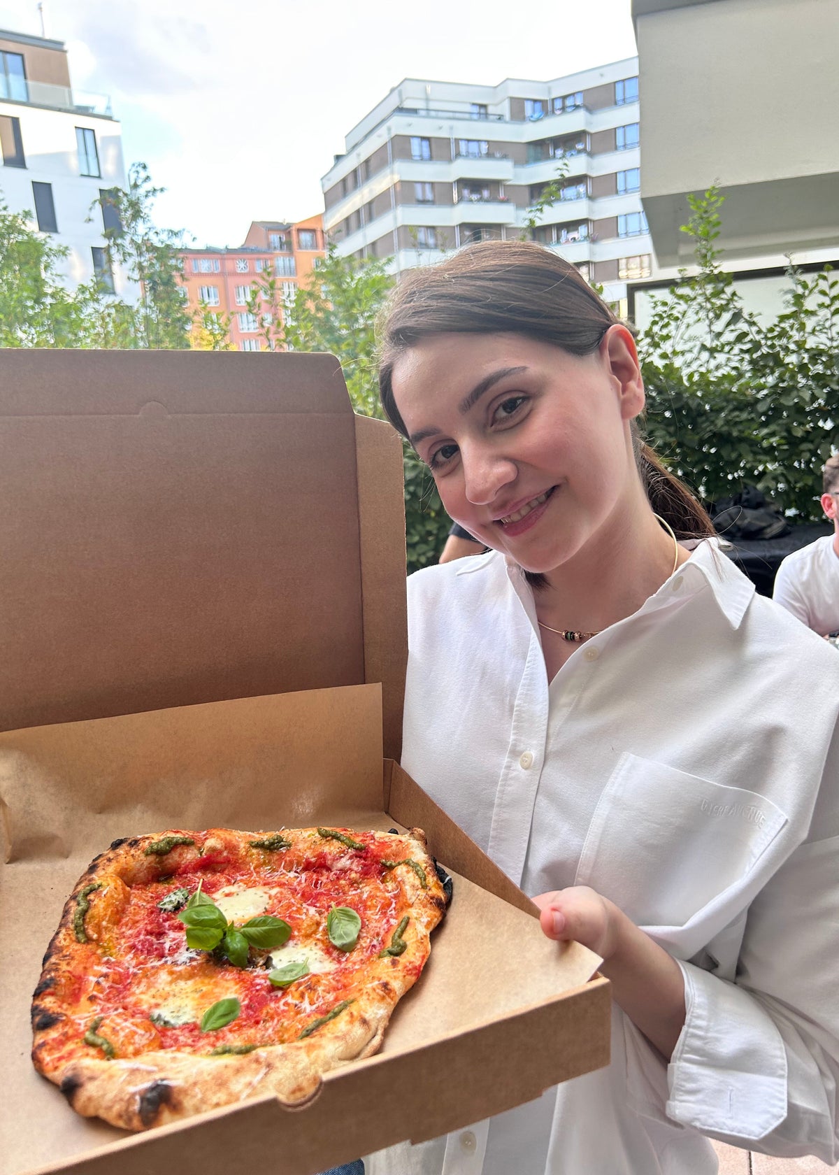 A woman in a white button-up shirt holds an open cardboard pizza box containing a freshly baked square pizza topped with tomato sauce, cheese, and green basil leaves. The background shows an outdoor patio setting with modern apartment buildings and greenery.
