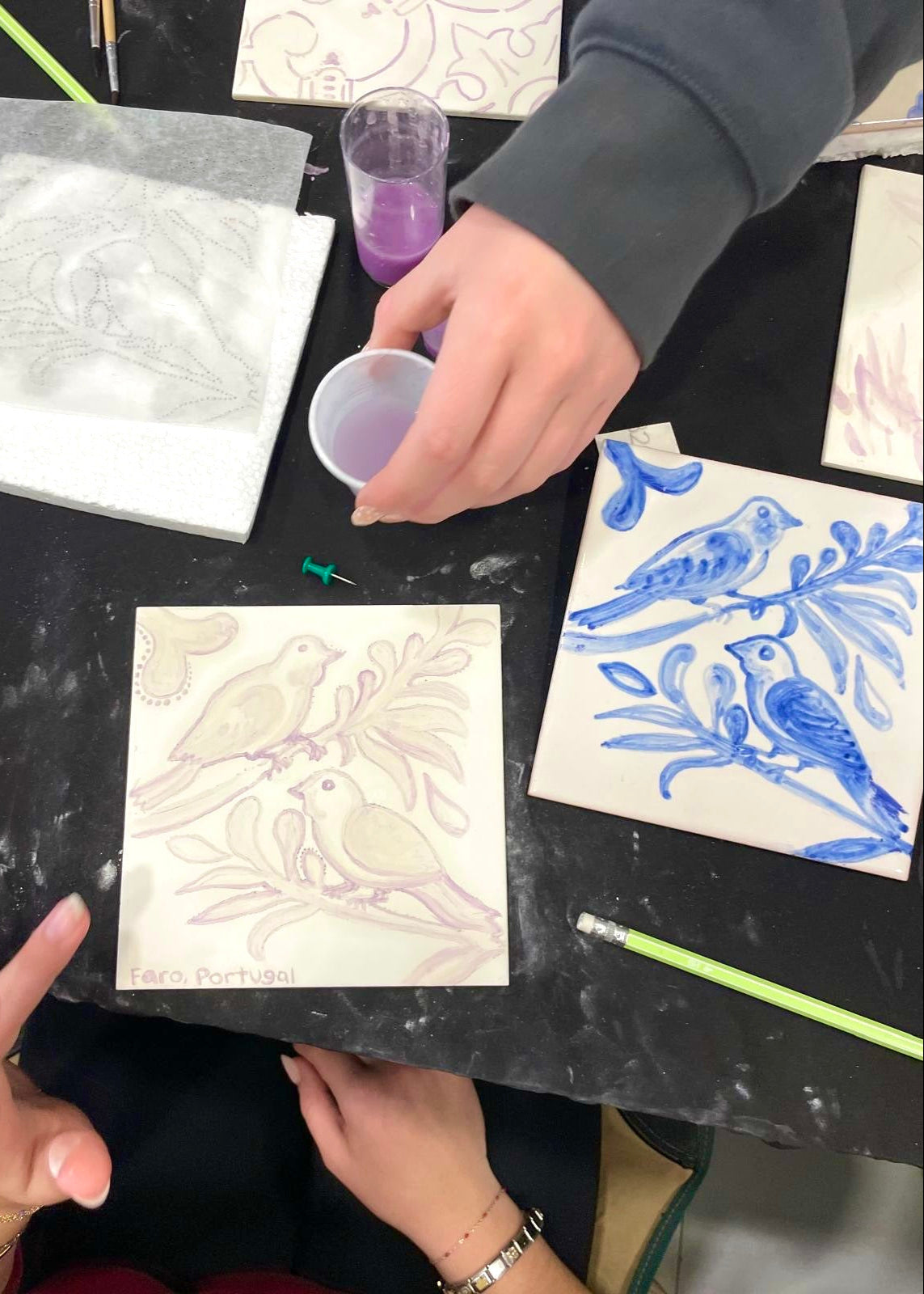 Person working on a ceramic tile with blue bird design and another with purple outline on a black surface.