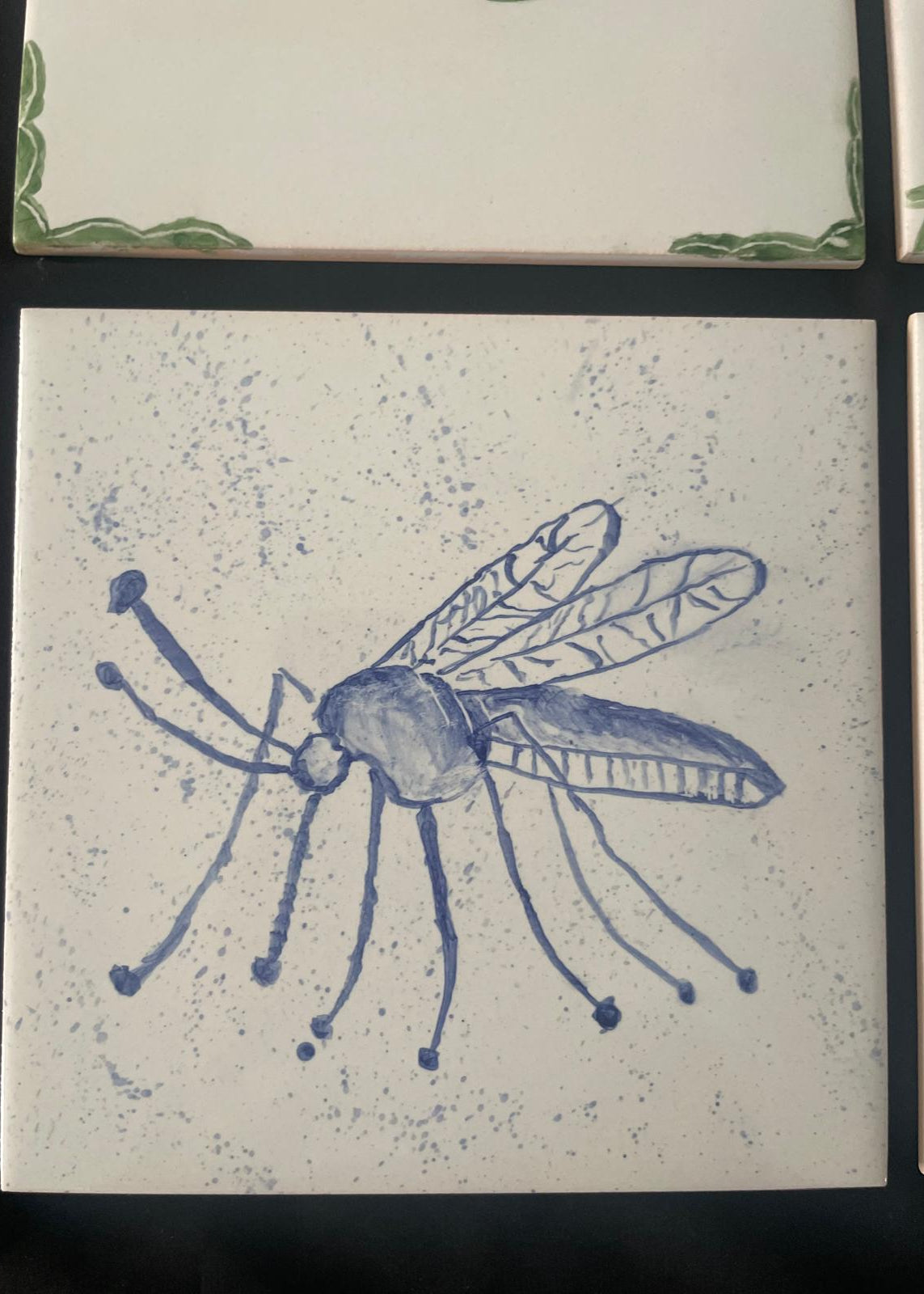 A square ceramic tile featuring a blue hand-painted illustration of a mosquito on a speckled off-white background, bordered by a dark surface.